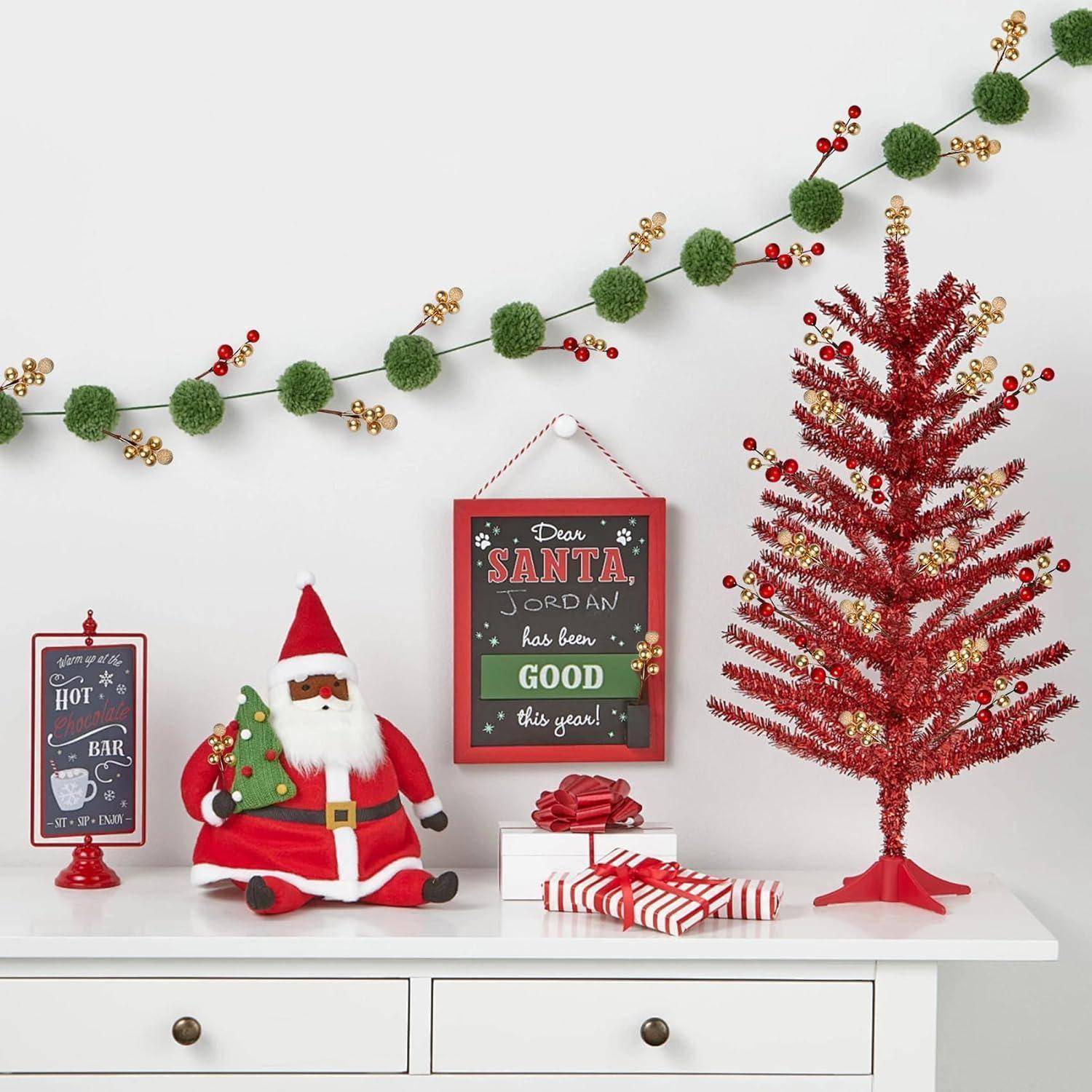 Eine festliche Weihnachtsdekoration mit einer Weihnachtsmann-Figur, einem roten Lametta-Baum und Feiertagsschmuck auf einem weißen Tisch. Das Schild liest: „Lieber Weihnachtsmann, Jordan war dieses Jahr brav!