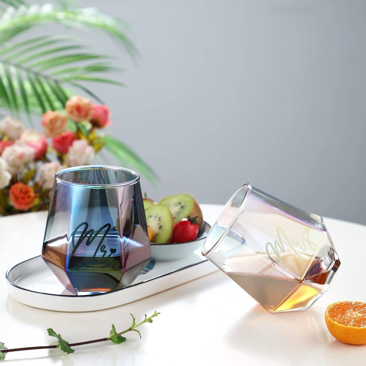 Zwei geometrische Glasbecher, einer aufrecht und einer geneigt, auf einem Tisch mit Früchten und Blumen im Hintergrund.