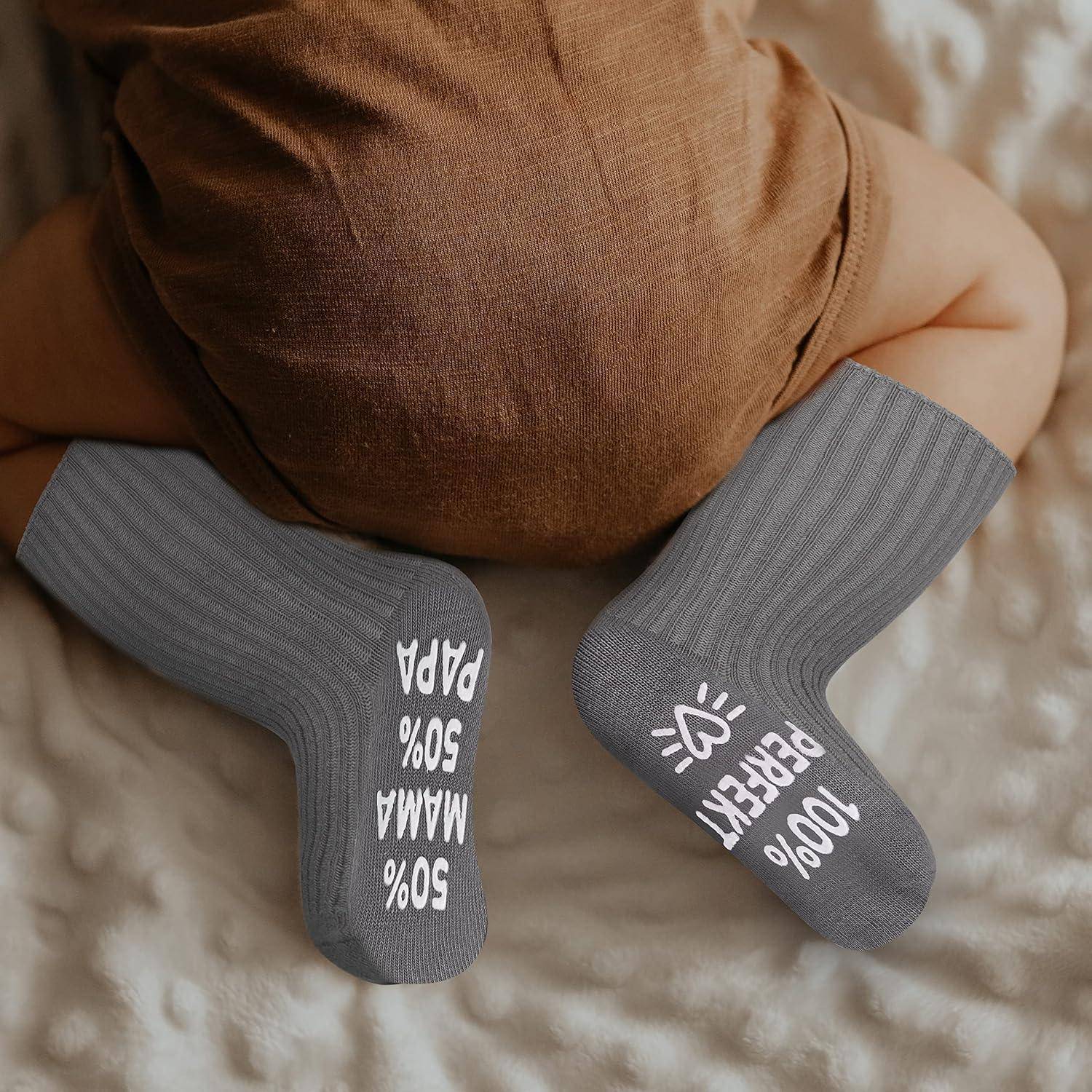 Baby liegt auf dem Bauch und trägt graue Socken mit weißem Text: '50% Mama, 50% Papa' auf einem Fuß, '100% Perfekt' auf dem anderen.