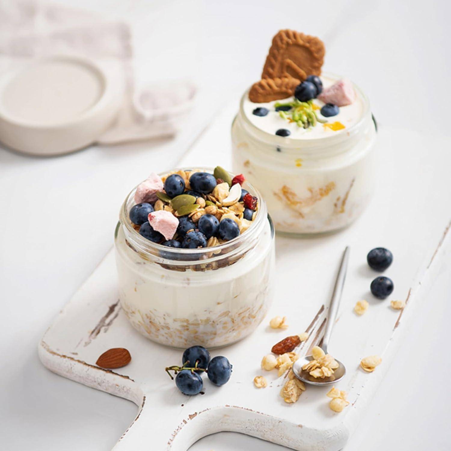 Zwei Gläser Joghurt, belegt mit Blaubeeren, Granola, Marshmallows und einem Keks auf einem weißen Brett. Ein Löffel und verstreute Nüsse befinden sich in der Nähe.