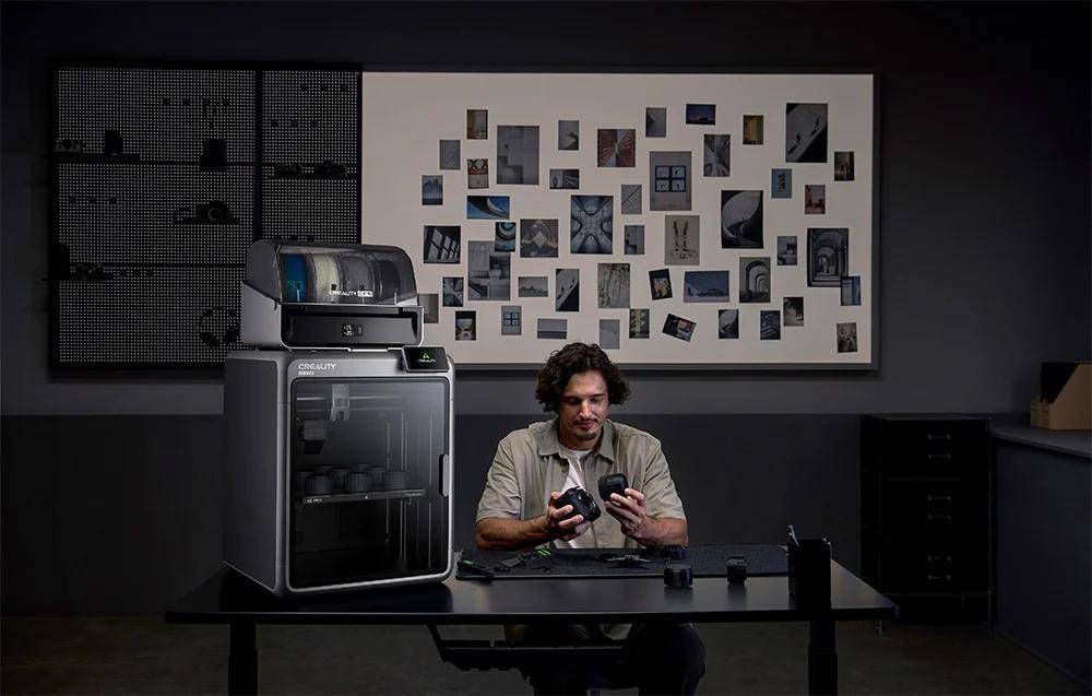 Eine Person sitzt an einem Tisch mit 3D-gedruckten Objekten, während ein 3D-Drucker und eine Wandanzeige mit Fotos und Designs in der Nähe stehen.