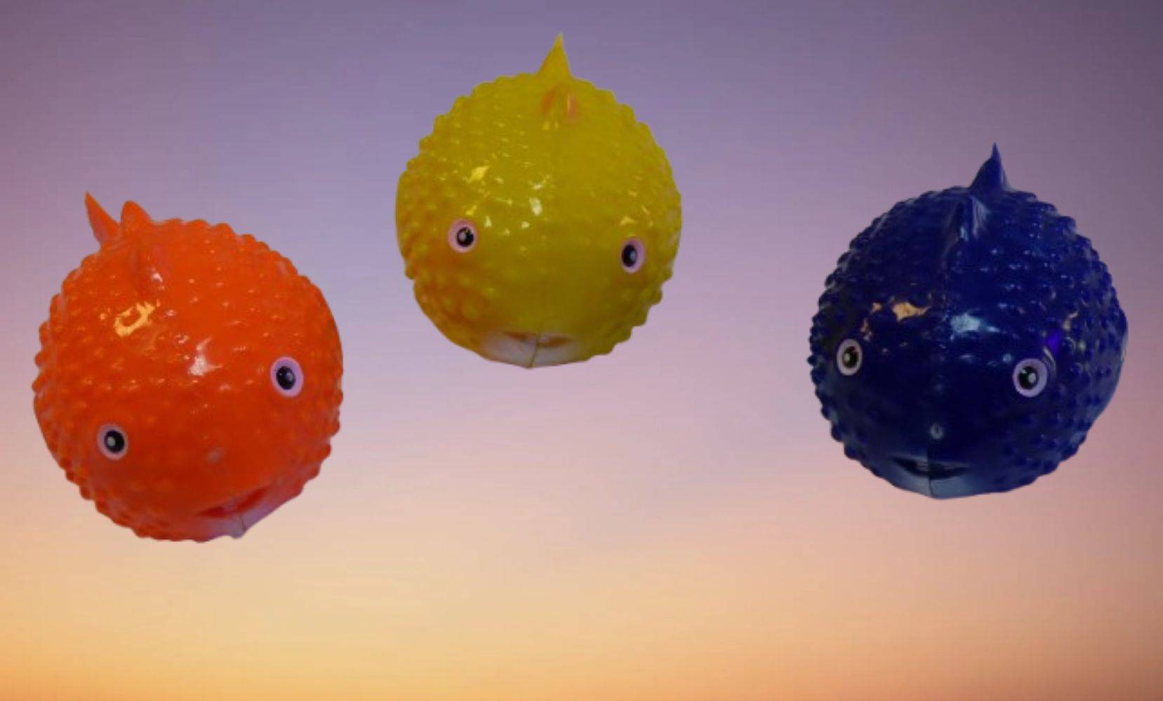 Drei farbenfrohe, fischförmige Luftballons schweben vor einem Sonnenuntergangshimmel, jeweils in Orange, Gelb und Blau, und erzeugen eine verspielt wirkende Szene.