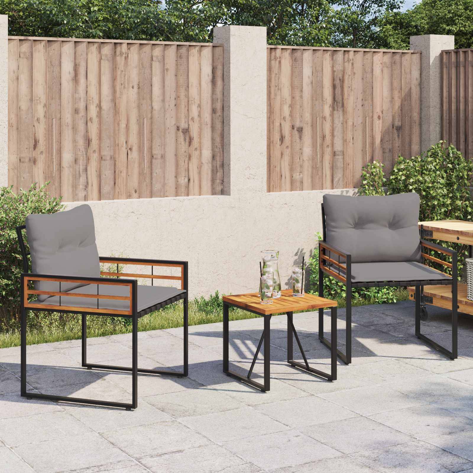 Terrasse mit zwei grauen gepolsterten Stühlen und einem kleinen Holztisch auf einer Betonoberfläche, umgeben von einem Holzzaun und Grünpflanzen im Hintergrund.