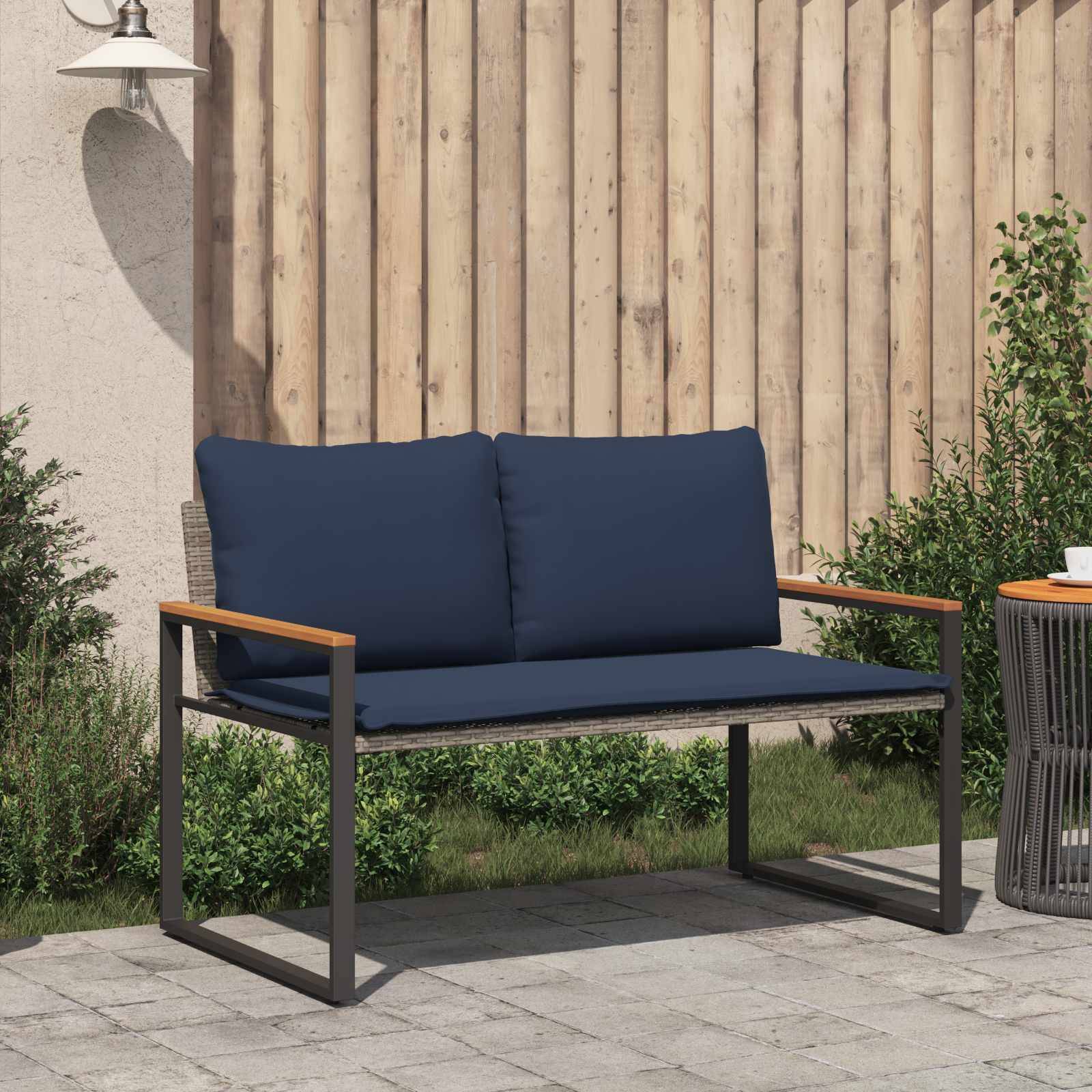 Eine Terrasse mit einer blau gepolsterten Bank und einem kleinen Outdoor-Tisch auf Steinplatten, umgeben von grünen Pflanzen, unter einer hellen Lampe.