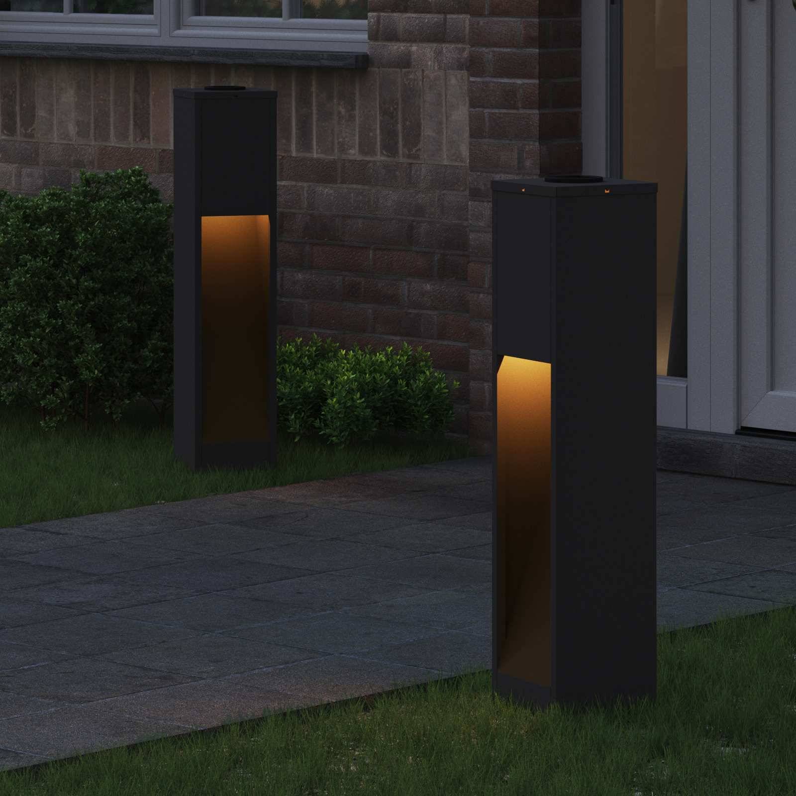Ein Gehweg, der zu einem Haus führt, wird von zwei modernen, rechteckigen Außenleuchten beleuchtet. Eine Hecke verläuft entlang der Hauswand.