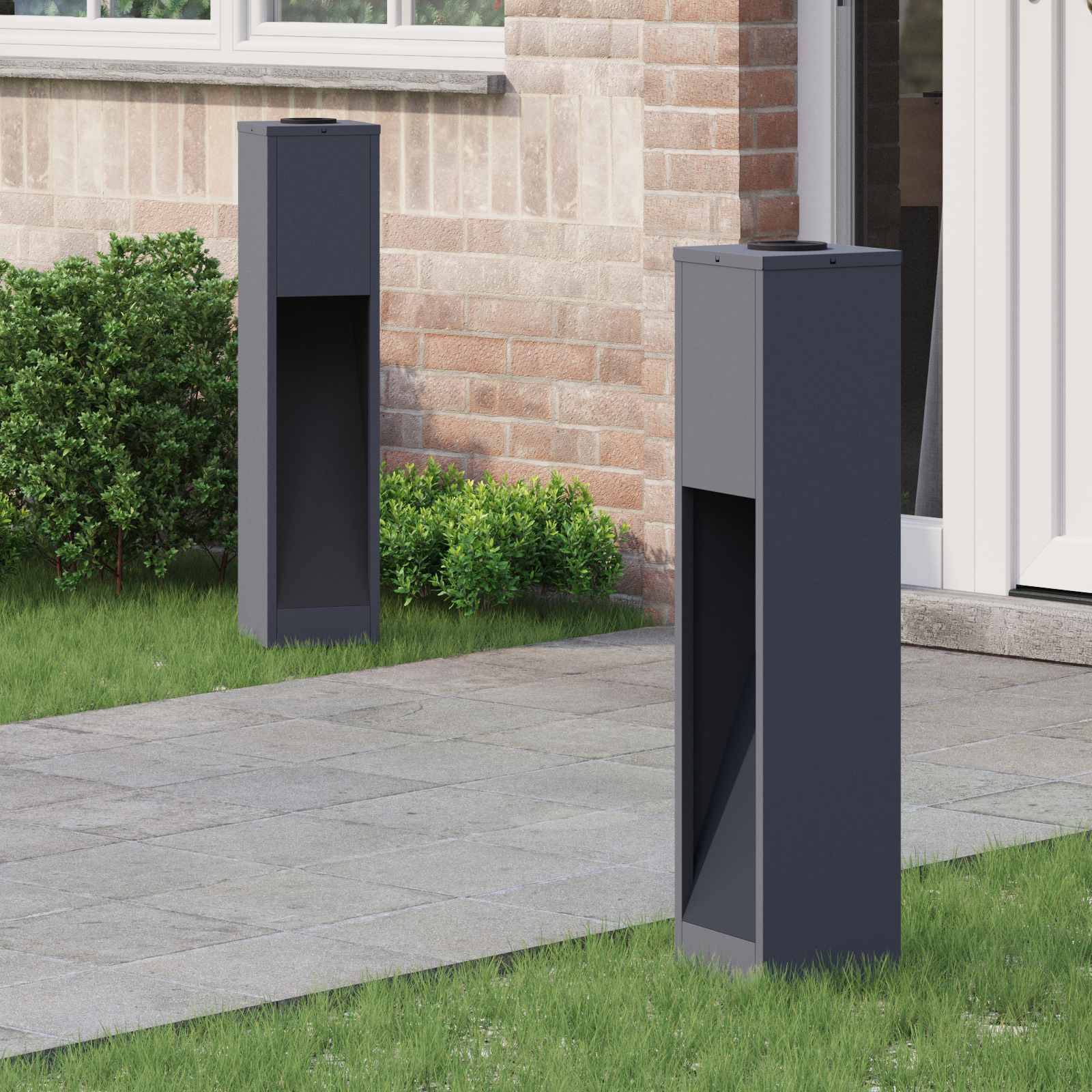 Zwei moderne dunkelgraue Paketboxen stehen auf einem gepflasterten Gehweg vor einem Ziegelhaus, umgeben von Gras und kleinen Sträuchern.