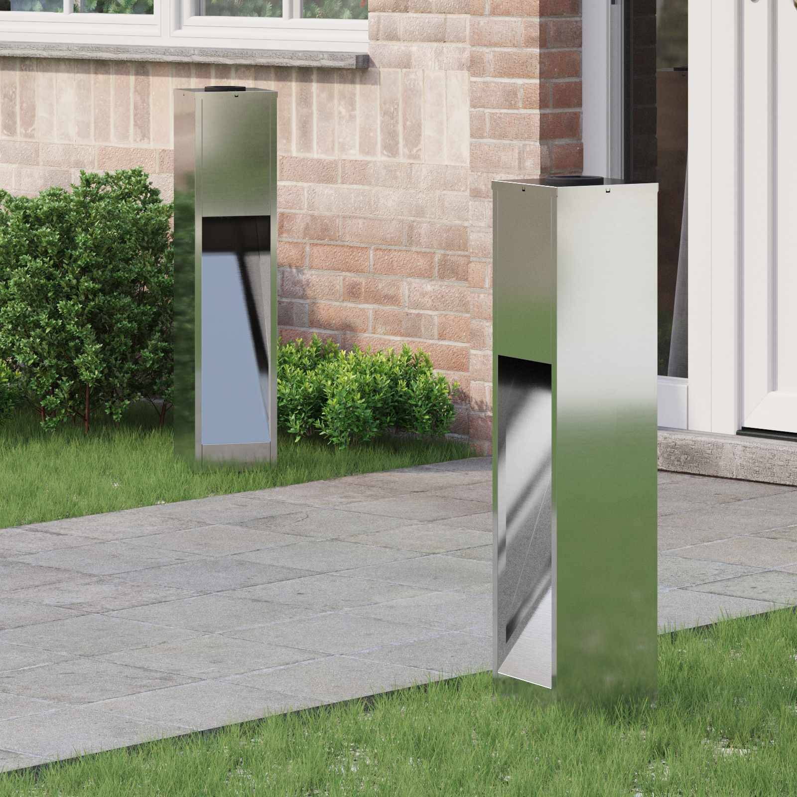 Zwei Edelstahl-Poller-Leuchten in einem Grasbeet neben einem Ziegelgebäude, die den Weg neben einem Gehsteig beleuchten.