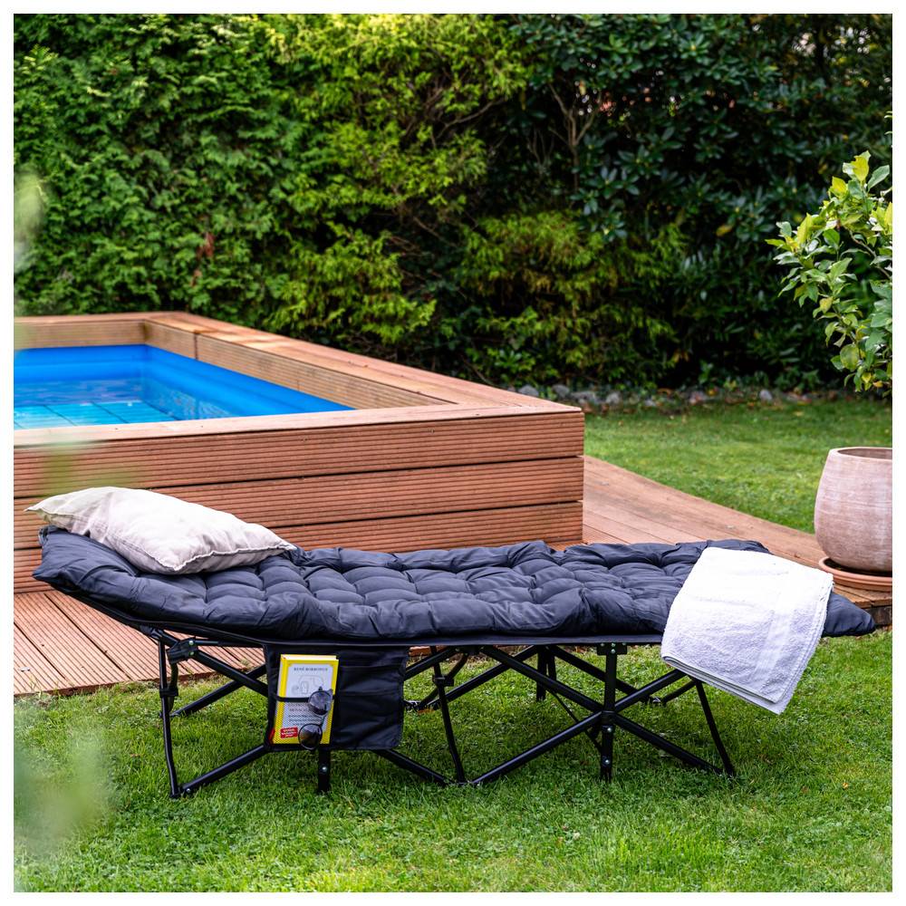 Ein tragbarer schwarzer Liegestuhl mit einem Kissen und einem Handtuch, der neben einem kleinen, quadratischen Pool platziert ist, umgeben von Gras und Bäumen.