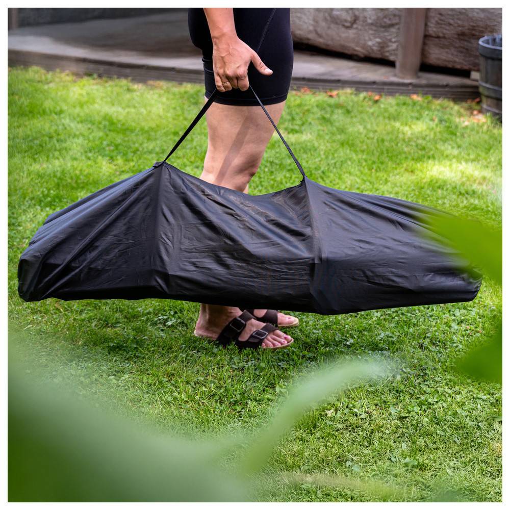 Person mit einer zusammengefalteten schwarzen tragbaren Hängematte in einer Tasche, die sich über eine Rasenfläche im Freien bewegt.