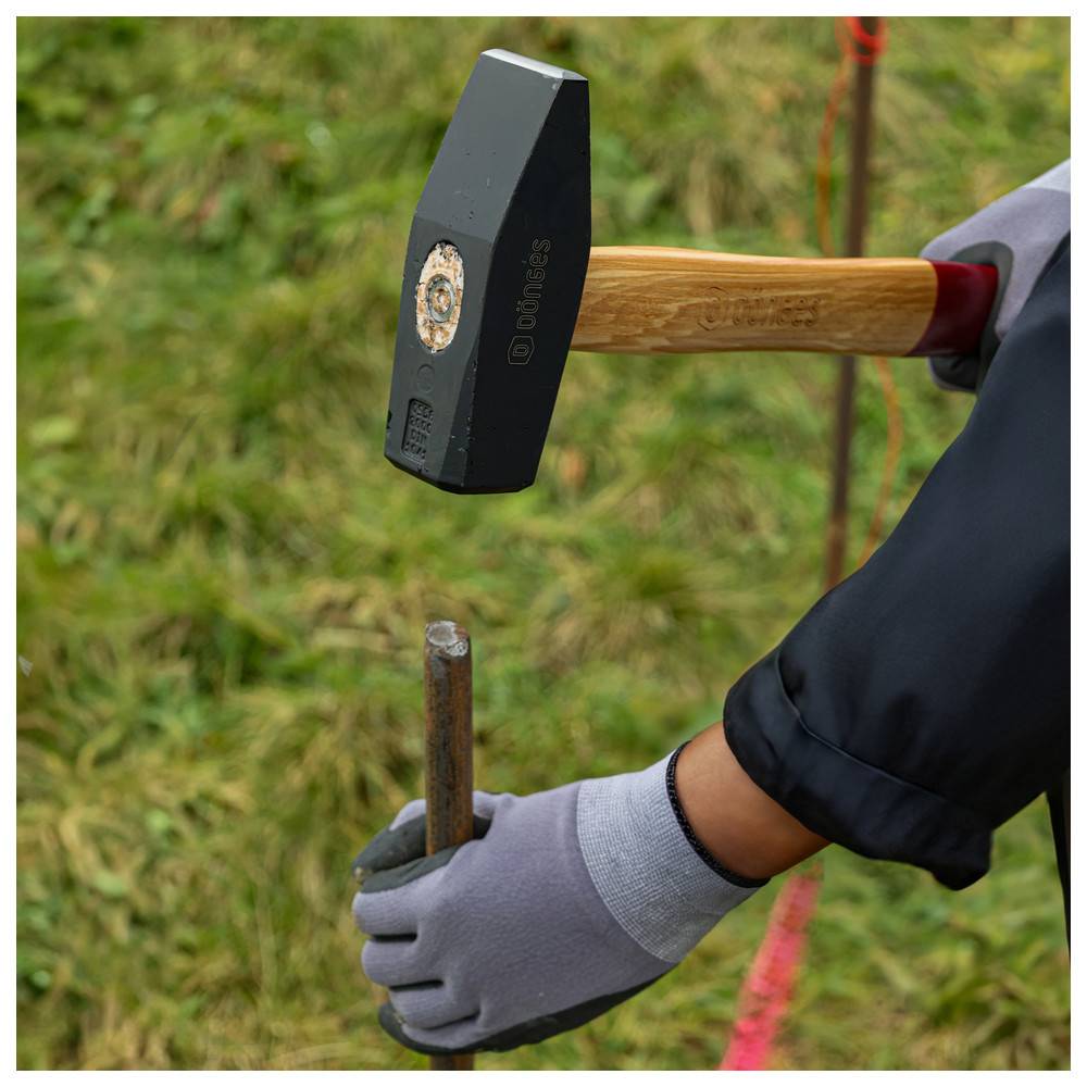 Eine Handschuh tragende Hand umfasst einen Metallstab, während ein Hammer darüber schwebt, bereit zum Schlag. Die Szene spielt sich in einem Grasgebiet ab, das mit rosa Linien markiert ist.