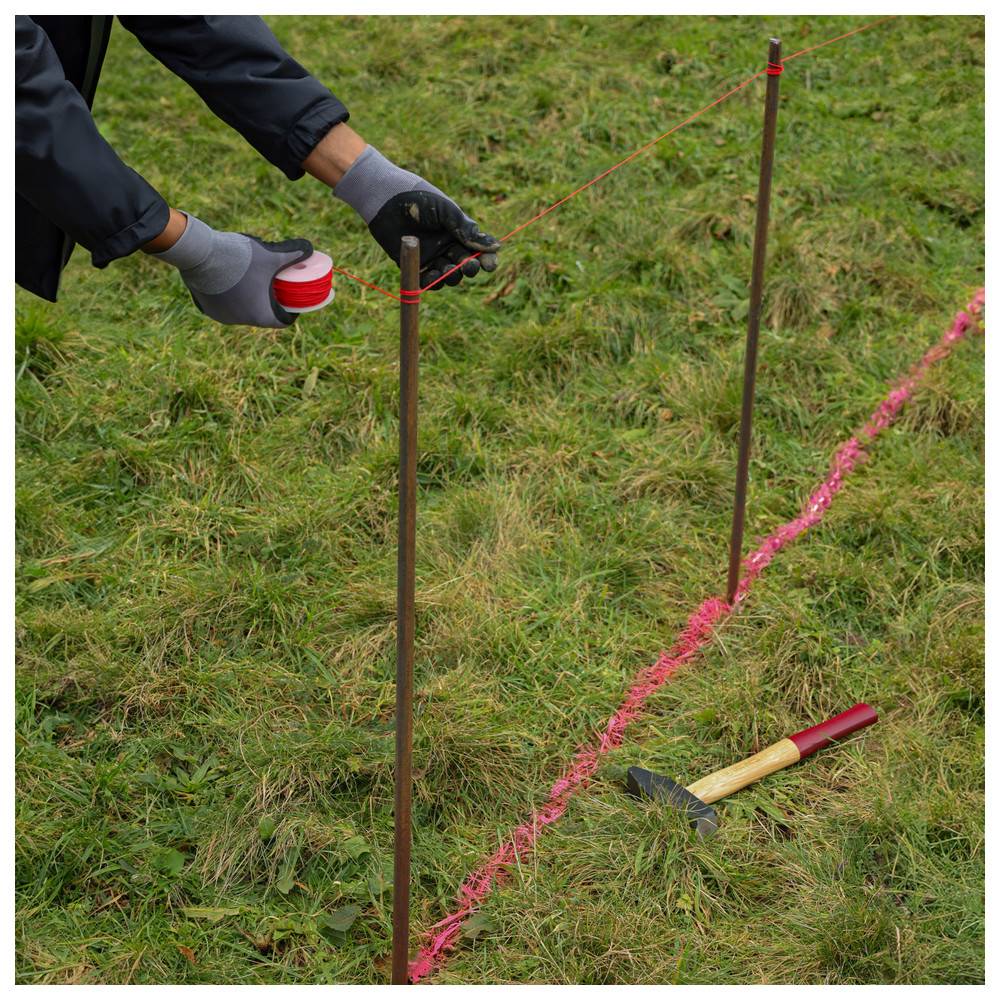 Eine Person mit Handschuhen bindet rote Schnur zwischen zwei Metallpfählen auf grässigem Boden, neben einem Hammer und einer Reihe von rosa Markierungen.