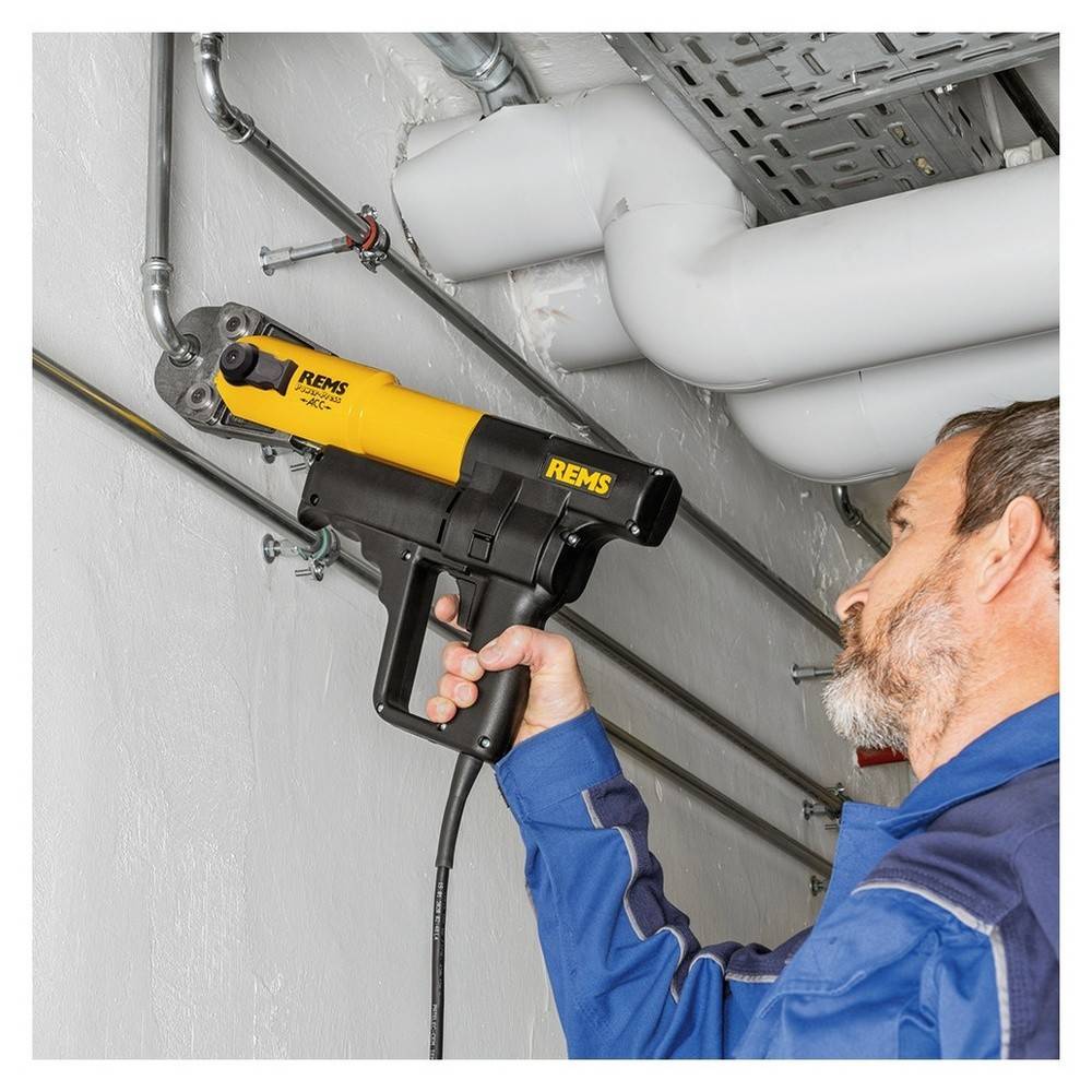 Eine Person verwendet einen elektrischen Rohrabschneider an Metallrohren entlang einer Wand. Das Werkzeug ist gelb und schwarz, wird fest umgriffen und zeigt präzise Arbeit.