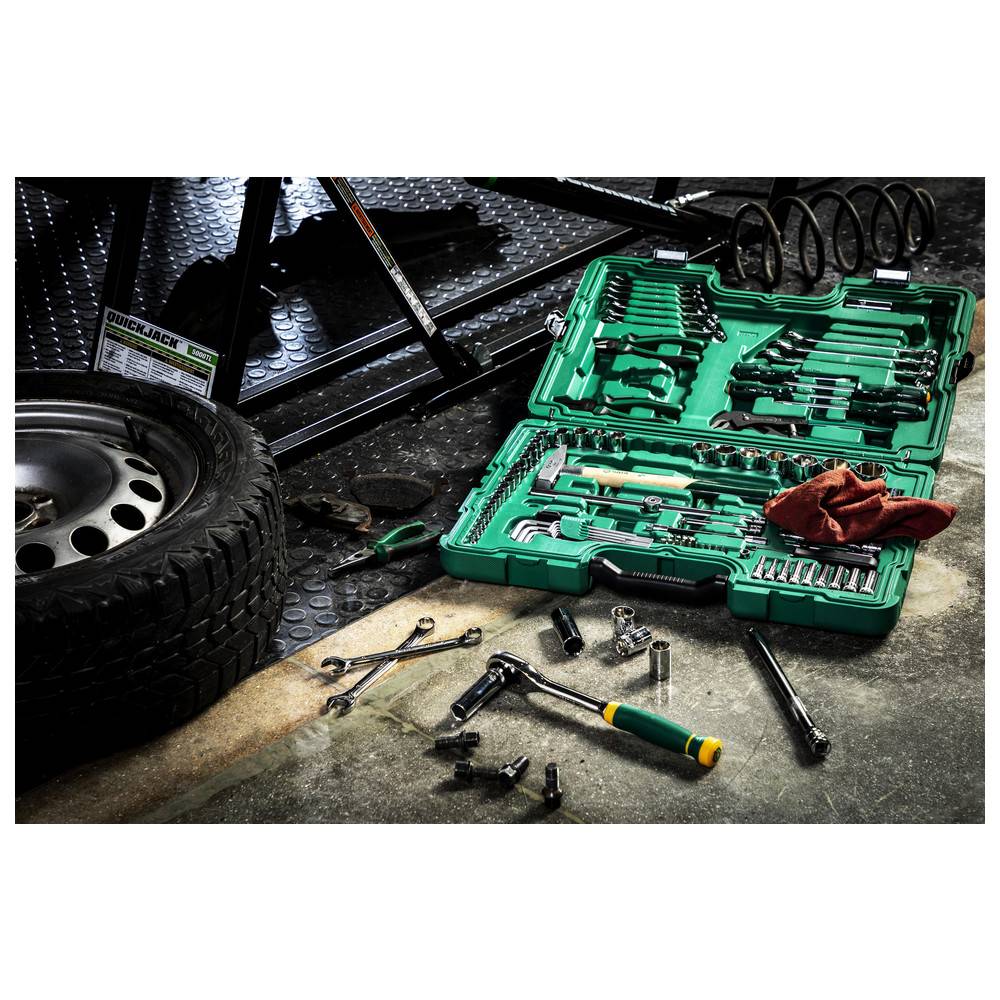 Ein grüner Werkzeugkasten mit verschiedenen Steckschlüsseln und Schraubenschlüsseln ist auf dem Boden neben einem Autoreifen und mechanischer Ausrüstung in einer Garagenumgebung geöffnet.