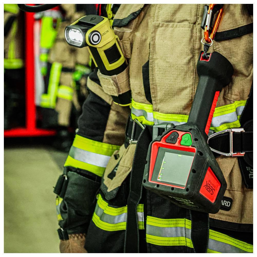 Ein Feuerwehrmann mit Schutzausrüstung hält zwei Geräte: eine an seiner Brust befestigte Taschenlampe und eine handgehaltene Wärmebildkamera.