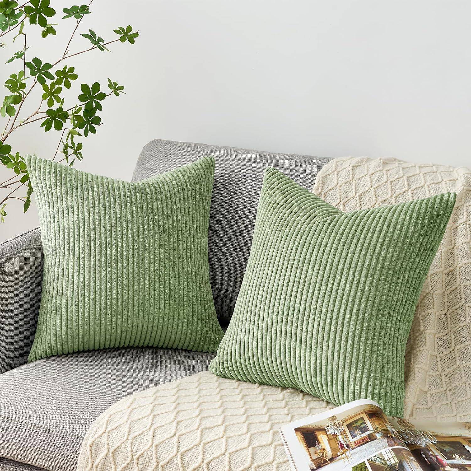 Grüne Kissen auf einem grauen Sofa mit einem strukturierten beigen Überwurf, neben einer Pflanze mit kleinen grünen Blättern, die eine gemütliche Wohnzimmeratmosphäre schaffen.
