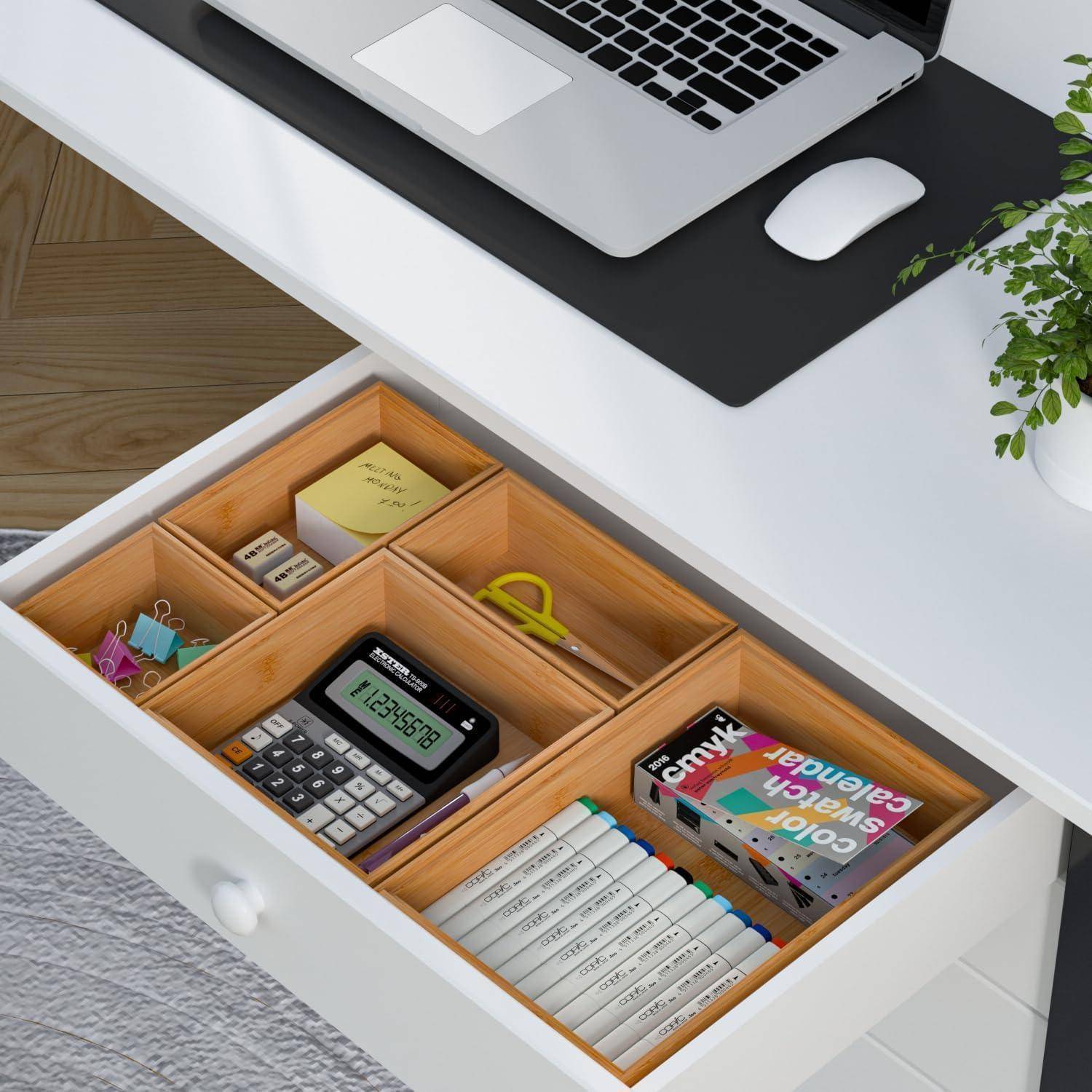 Eine offene Schublade in einem weißen Schreibtisch, organisiert mit Schreibwaren, einem Taschenrechner, Haftnotizen und Markern. Ein Laptop steht auf dem Schreibtisch darüber.