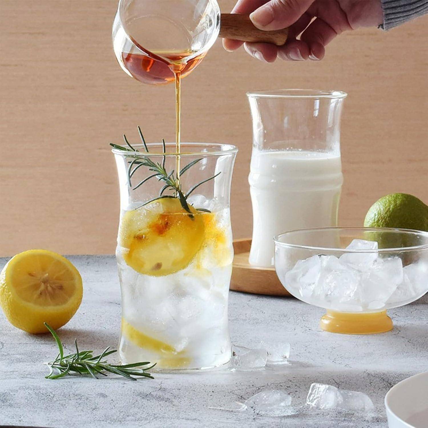 Eine Hand gießt Honig in ein Glas mit Eiszitronen-Wasser und Rosmarin. Ein Glas Milch, eine Limette und eine Schüssel mit Eis befinden sich im Hintergrund.