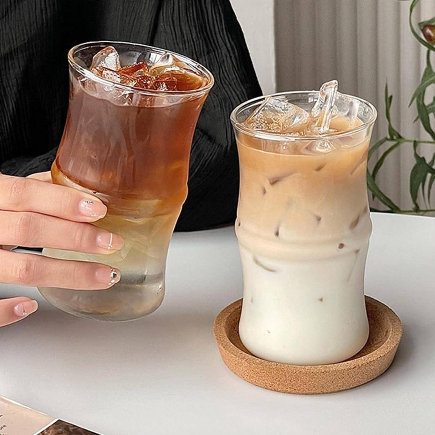 Eine Hand, die einen Eiskaffee hält, neben einem Glas mit Eismilchtee auf einem Untersetzer, beide auf einem Tisch mit einer Pflanze im Hintergrund.