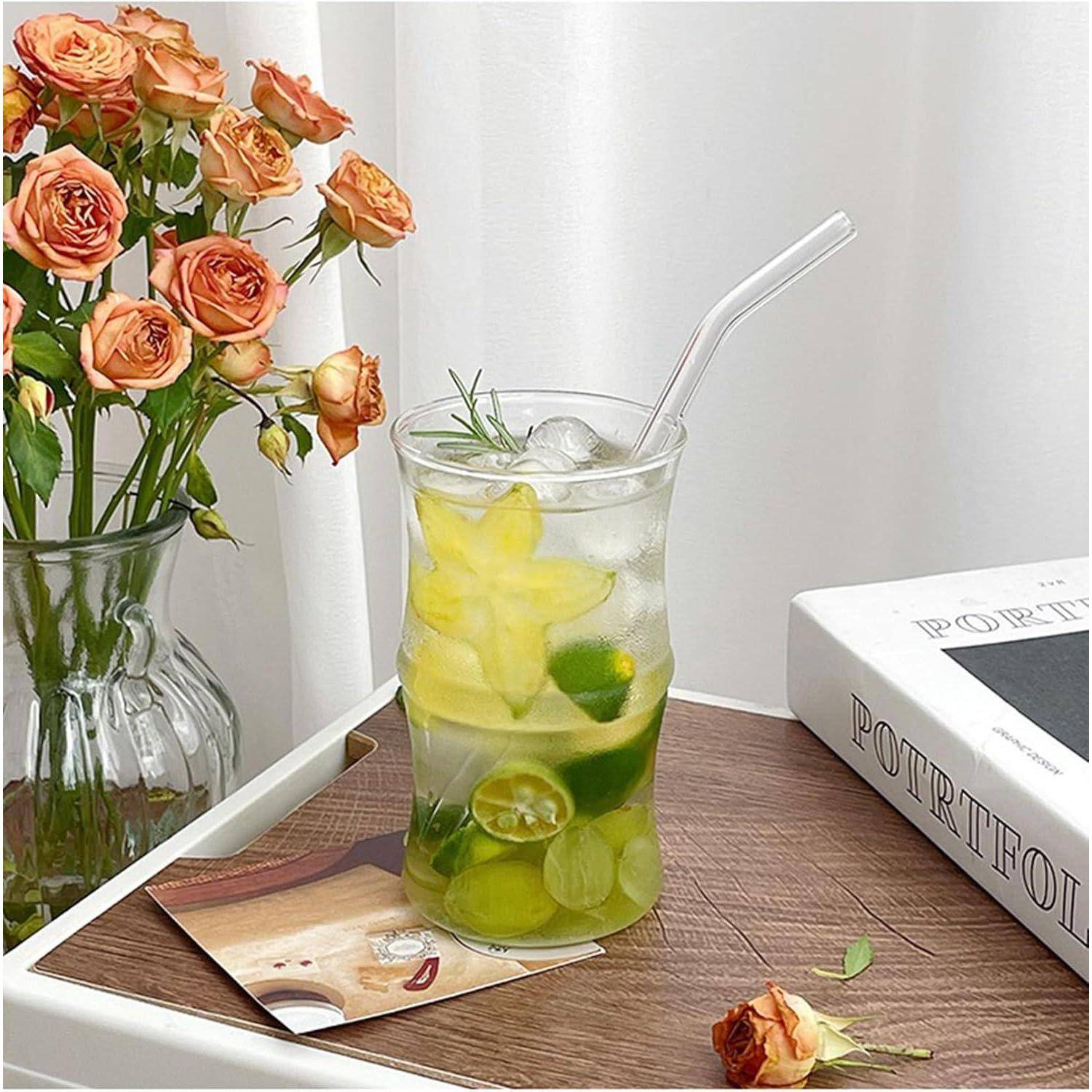 Ein Glas mit eisgekühltem Zitronen-Limetten-Getränk und einem Strohhalm auf einem Holztablett. Eine Vase mit orangefarbenen Rosen und ein Buch befinden sich im Hintergrund.
