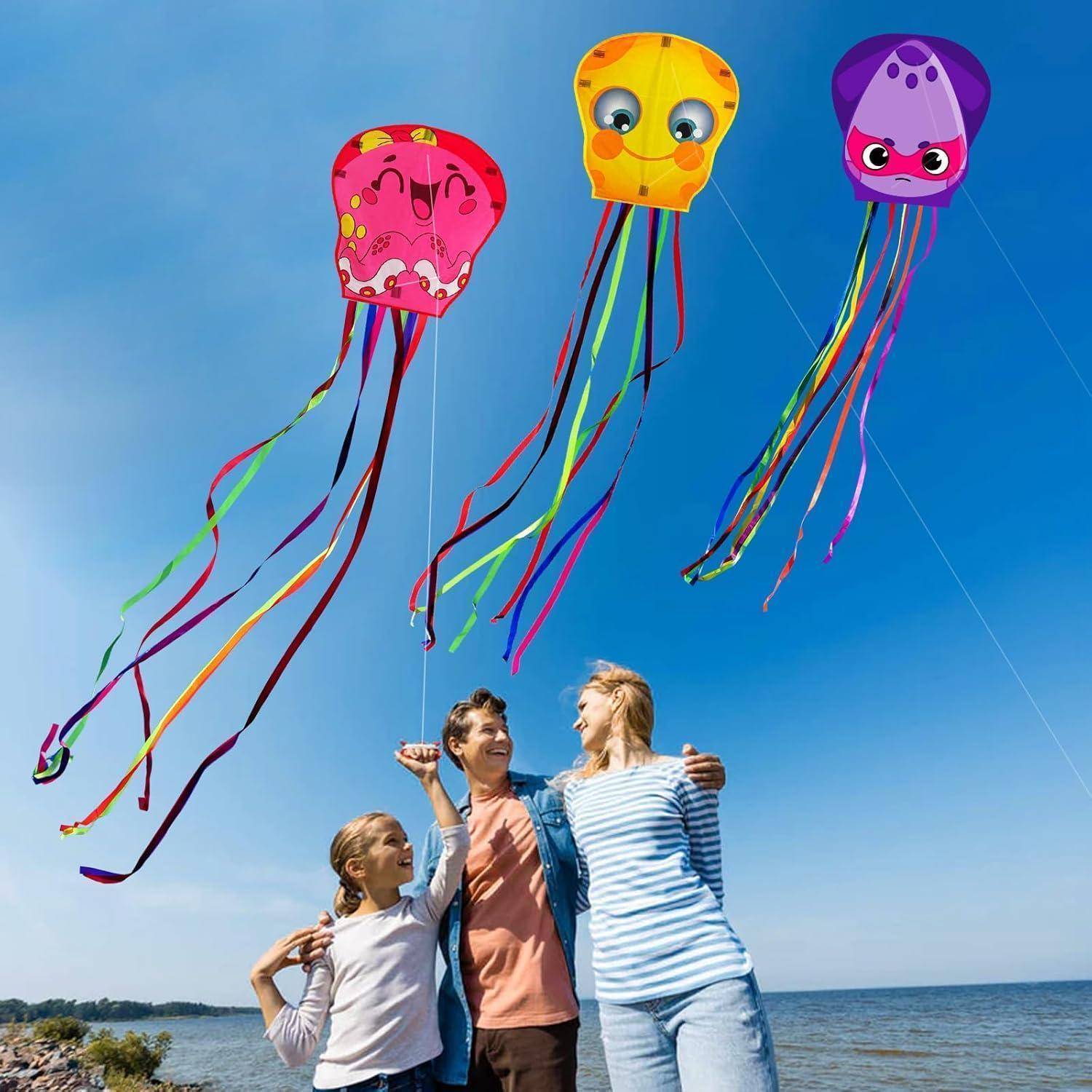 Eine Familie mit drei Personen lächelt, während sie bunte, quallen-förmige Drachen am Strand fliegen, mit einem klaren blauen Himmel im Hintergrund.