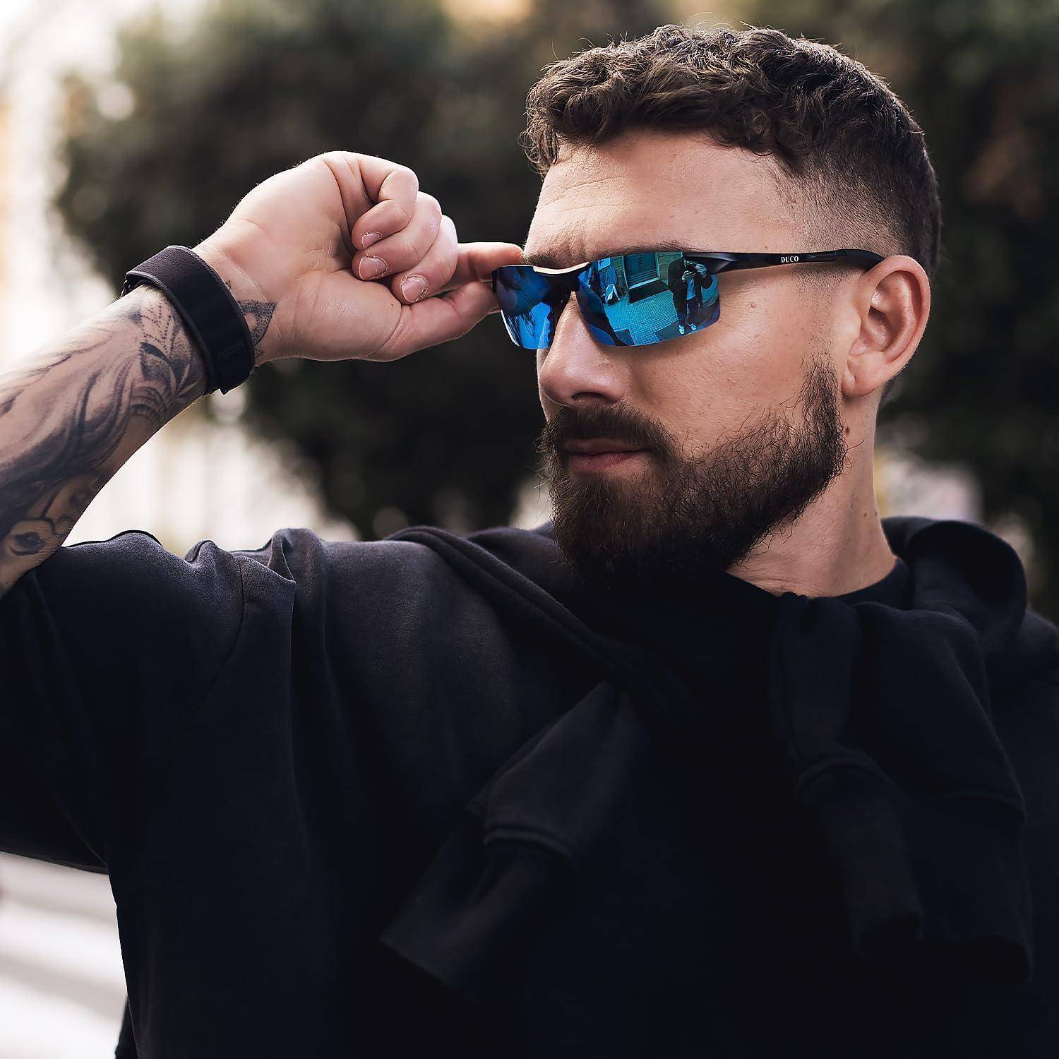 Ein Mann mit Bart und Sonnenbrille posiert im Freien und richtet seine Sonnenbrille her. Er hat eine Tätowierung auf dem Arm und trägt eine dunkle Jacke.
