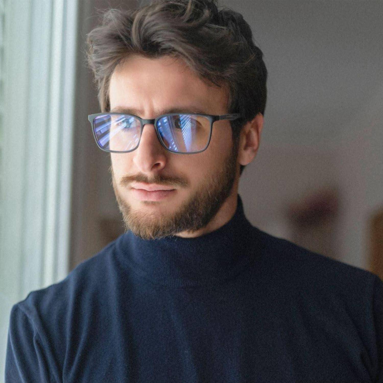 Eine Person mit Brille und dunklem Rollkragenpullover schaut aus einem Fenster mit Jalousien, wobei das Licht Reflexionen auf den Gläsern wirft.