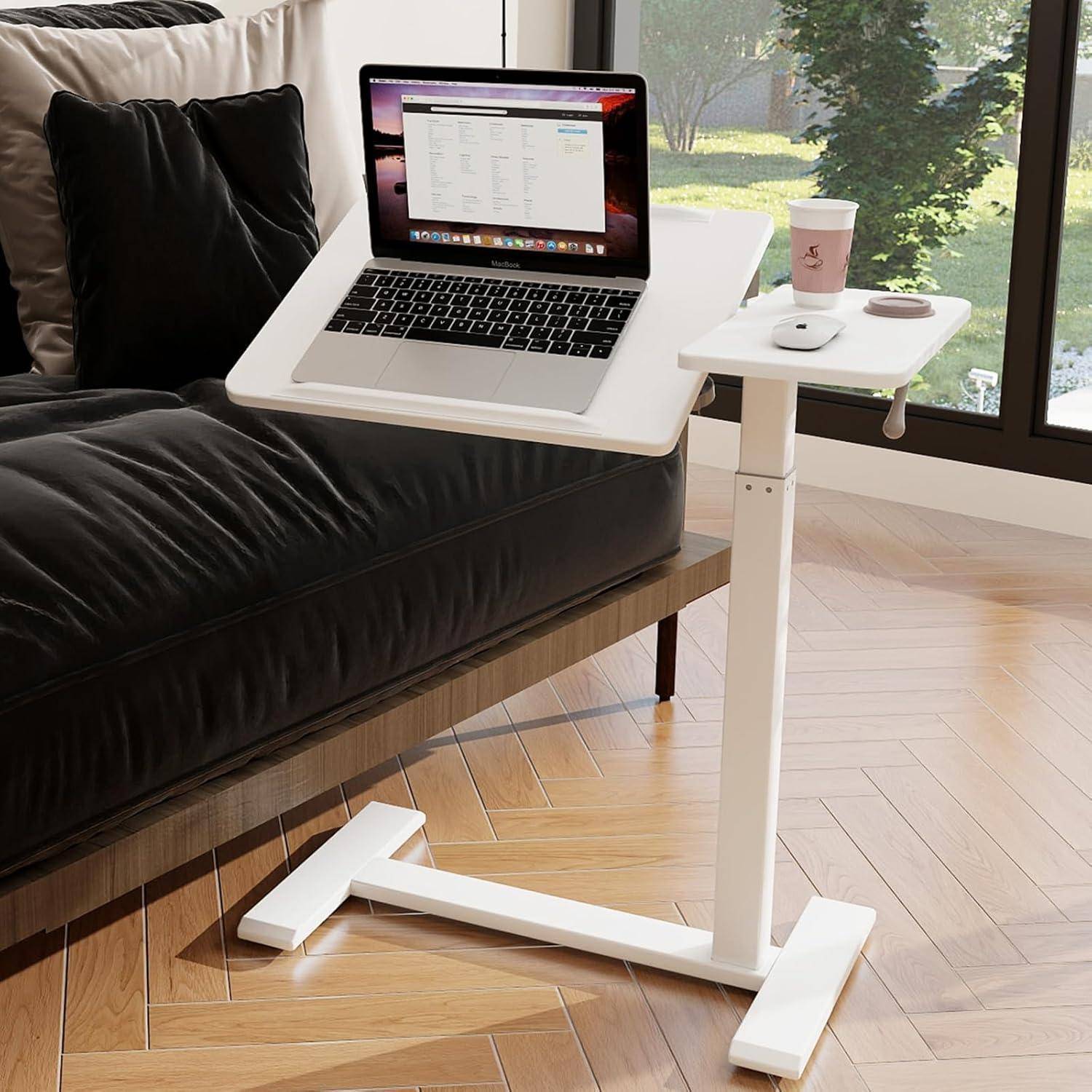 Ein Laptop mit geöffnetem Bildschirm steht auf einem kleinen, verstellbaren weißen Schreibtisch neben einem Sofa, mit Blick aus dem Fenster auf Bäume im Hintergrund.