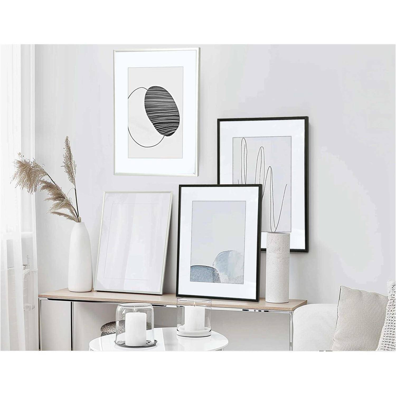 Ein minimalistischer Raum zeigt gerahmte abstrakte Kunst an einer weißen Wand, einen schmalen Konsolentisch mit einer weißen Vase, in der getrocknete Gräser stehen, und Kerzen.