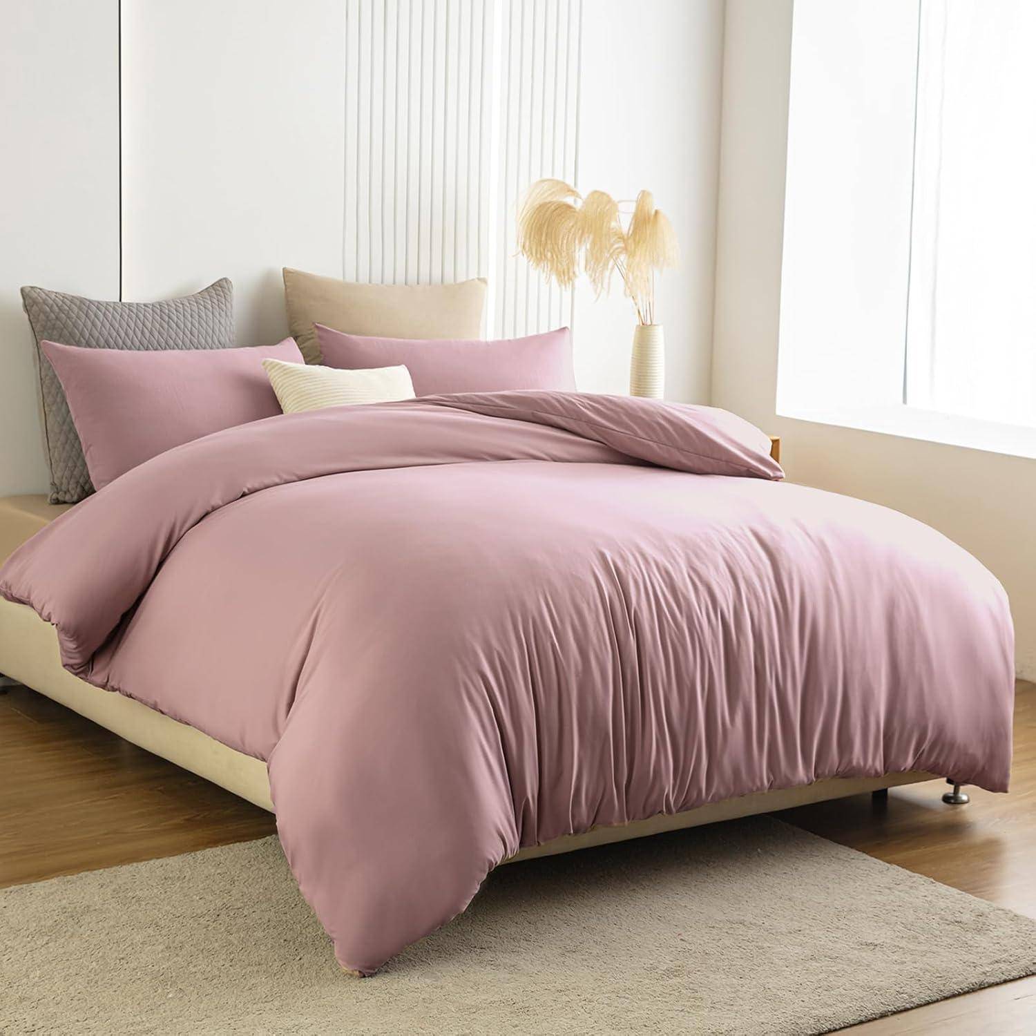 Ein ordentlich gemachtes Bett mit einer hellrosa Bettdecke und Kissen in weichen, neutralen Tönen. Eine Pflanze in einer Vase und transparente Gardinen schmücken den Hintergrund.
