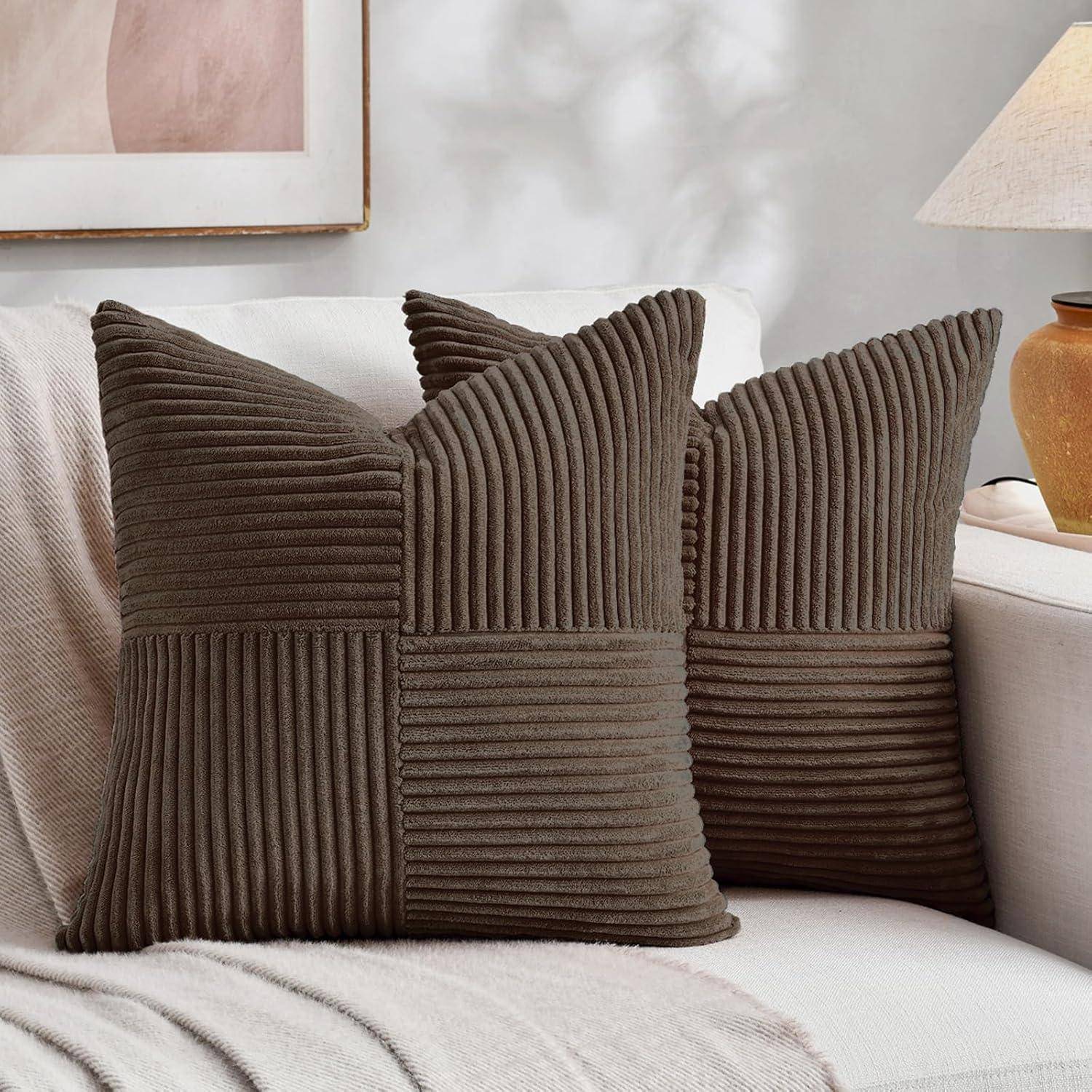 Zwei braune quadratische Kissen mit gerippter Textur auf einem beigen Sofa neben einer Tischlampe; gerahmte Kunstwerke teilweise im Hintergrund sichtbar.