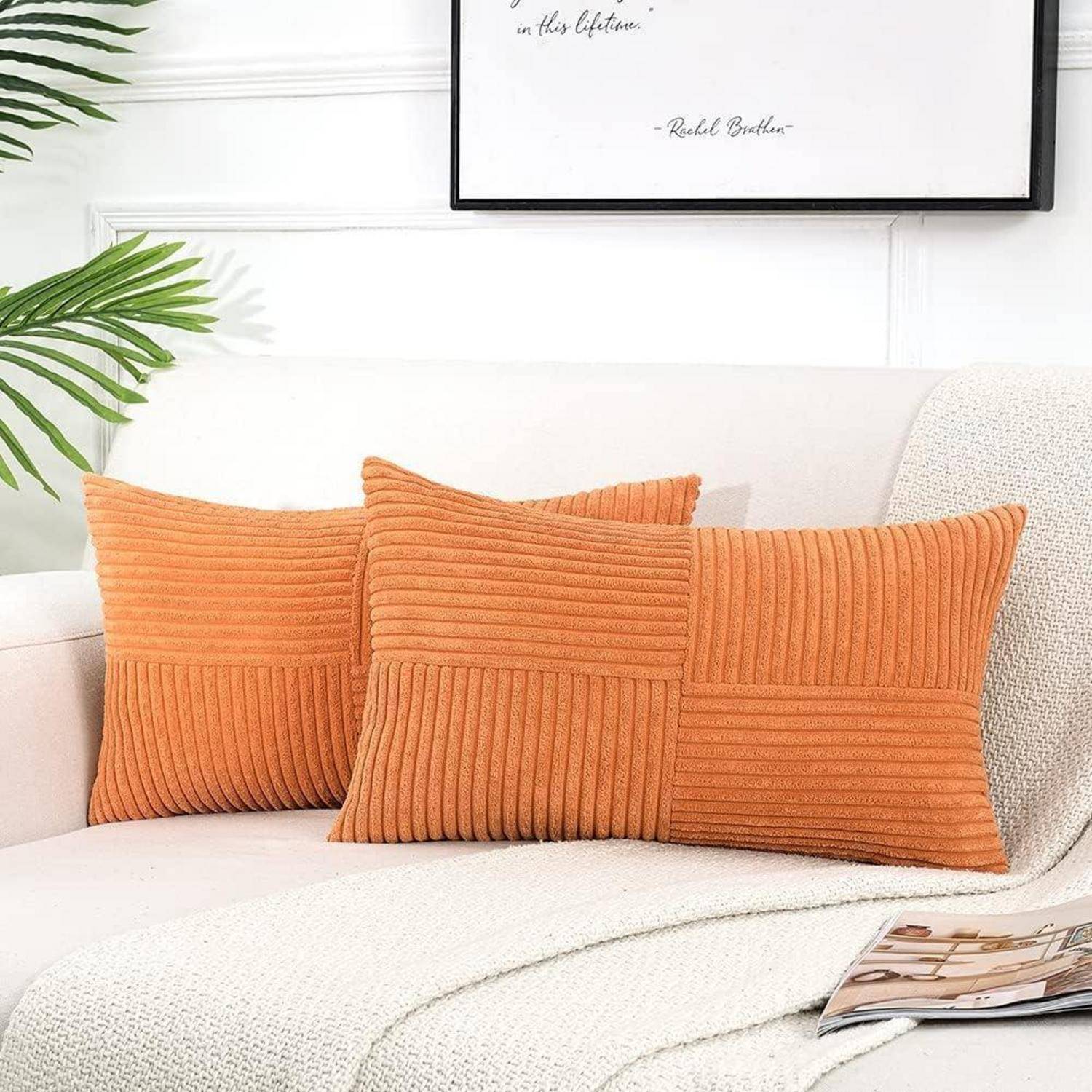 Orangefarbene gerippte Kissen auf einem cremefarbenen Sofa, neben einer Zimmerpflanze im Tropendesign. Ein gerahmtes Zitat hängt darüber, teilweise verdeckt. Eine Zeitschrift liegt auf dem Sofa.