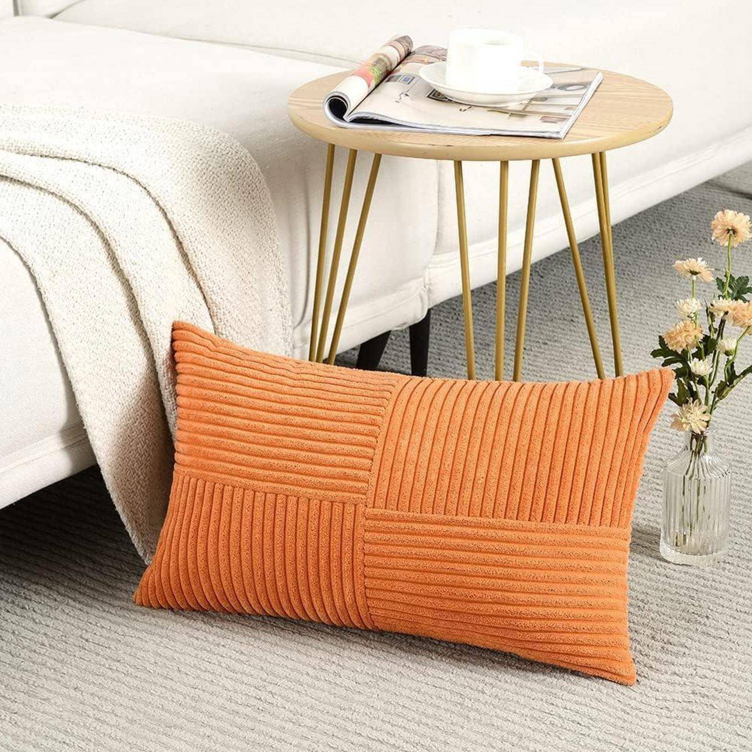 Ein orangefarbenes Zierkissen mit gerippter Textur liegt auf einem hellen Teppich neben einem Holz-Beistelltisch, auf dem eine Zeitschrift und eine Couch stehen.