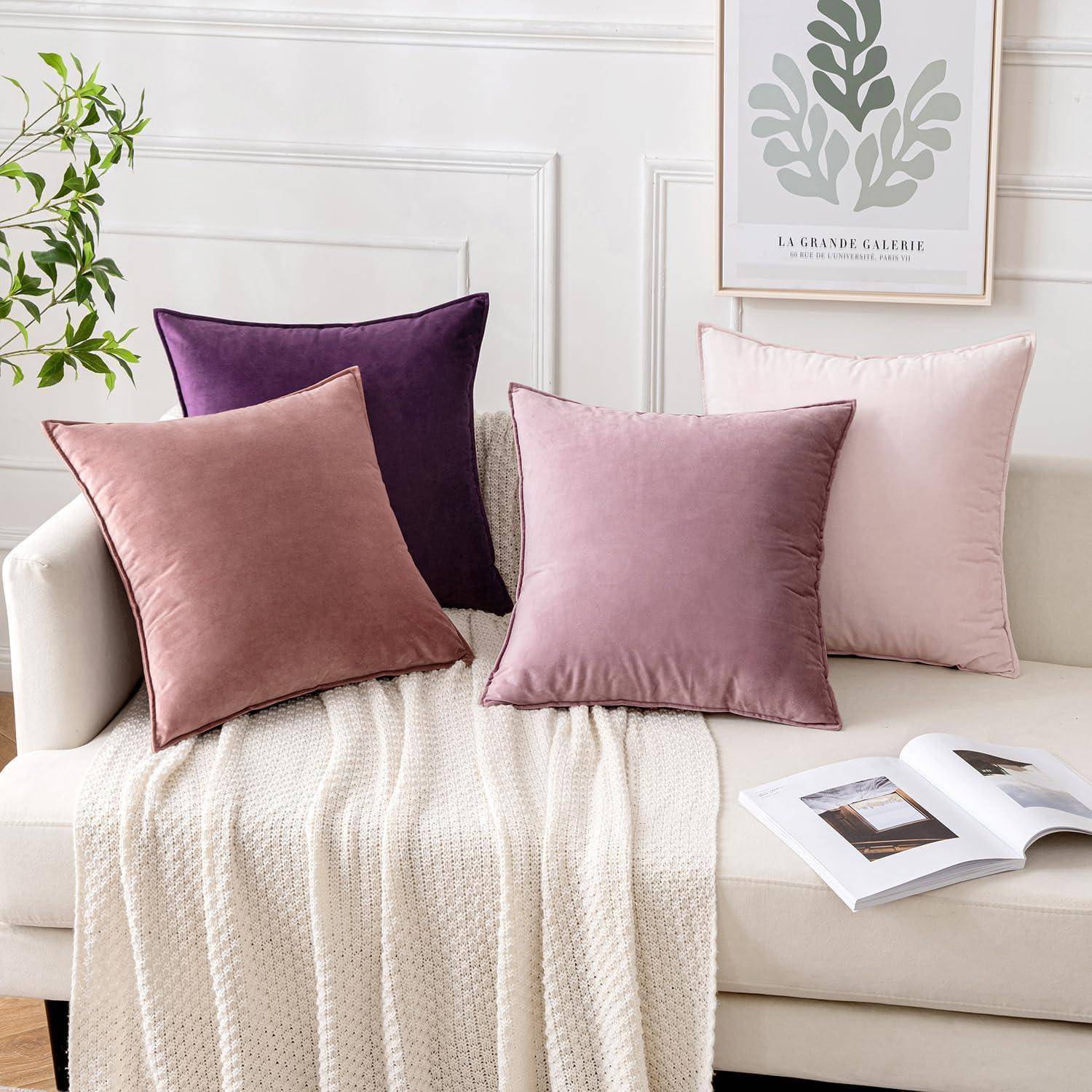 Eine weiße Couch mit einer beigen Strickdecke und vier Samtkissen in Rosa- und Lilatönen. Ein gerahmter abstrakter Blätterdruck hängt darüber.