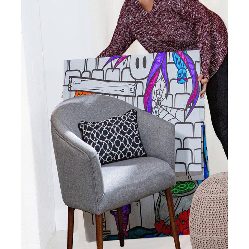 Eine Person in einem gemusterten Pullover, die neben einem großen, bunten Gemälde mit abstrakten Formen und einem grauen Stuhl steht.