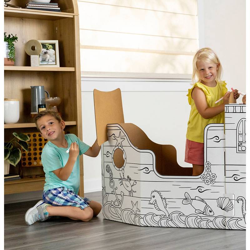 Zwei Kinder, eines stehend und eines sitzend, spielen mit einem großen Pappschiff-Spielzeug, das mit Meeresszenen verziert ist, in einem Raum mit Holzregalen.