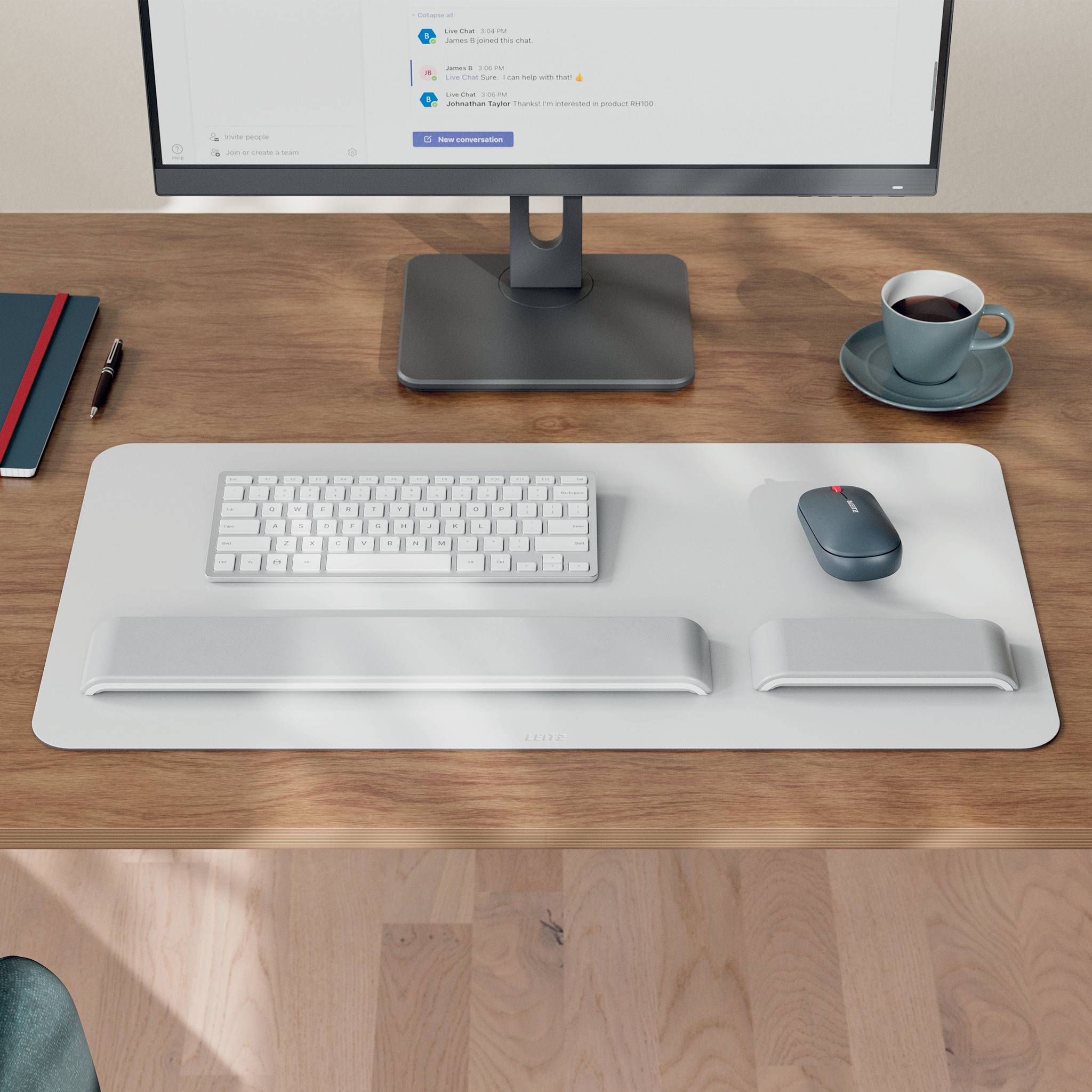 Minimalistischer Desktop-Arbeitsplatz mit Monitor, Tastatur, Maus, Handgelenkstützen, Kaffeetasse, Notizbuch und Stift auf einem Holzschreibtisch.