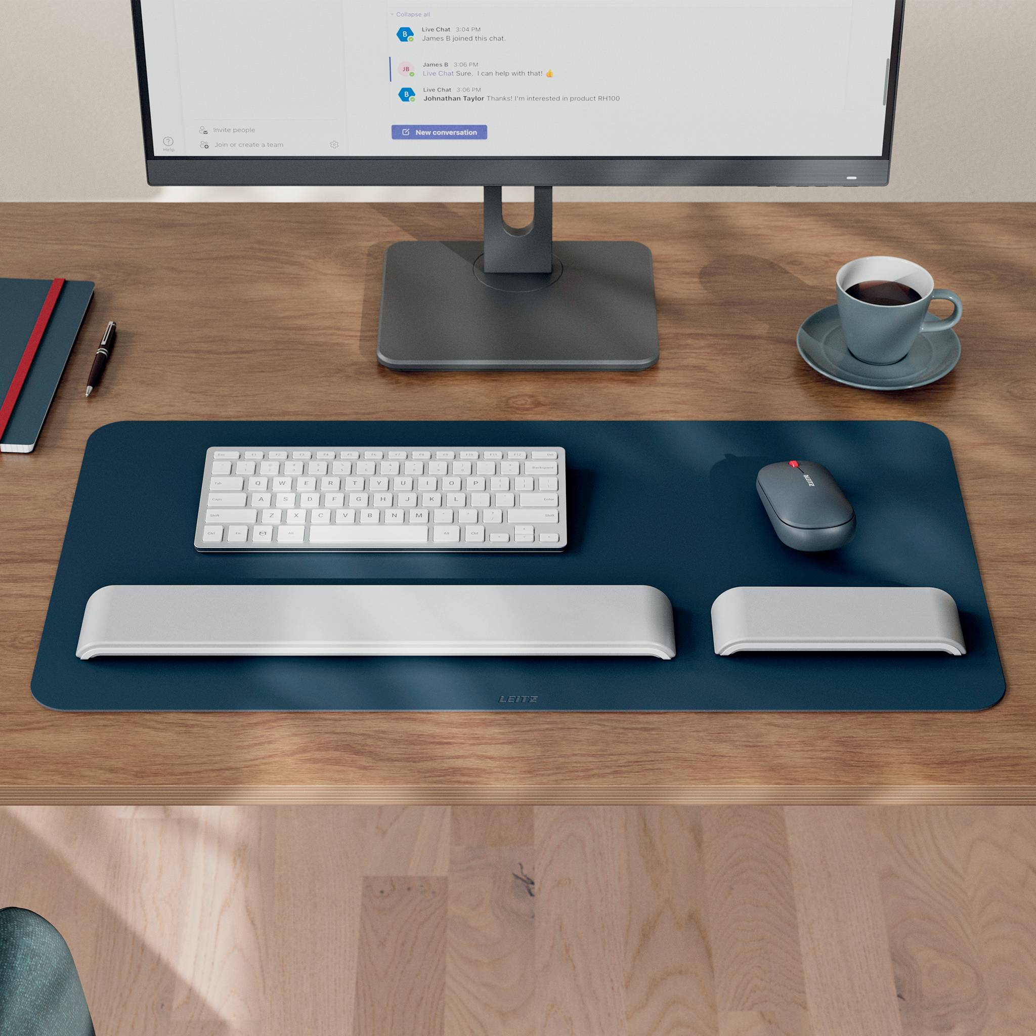 Minimalistische Schreibtischeinrichtung mit Monitor, Tastatur, Maus, Handgelenkstützen, Kaffeetasse, Notizbuch und Stift auf einem Holztisch.