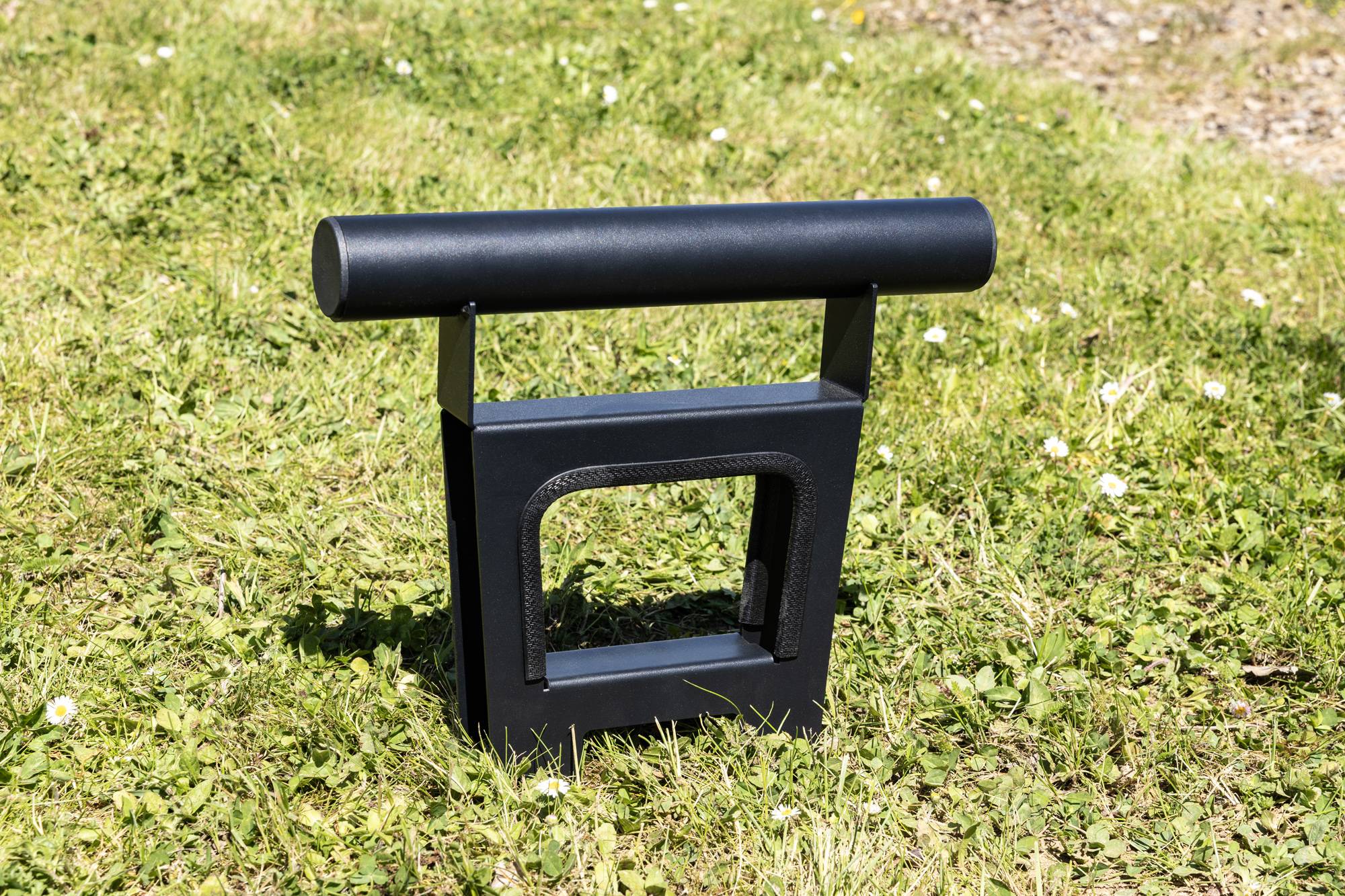 Eine schwarze Metallstruktur mit einer T-förmigen Spitze steht auf grünem Gras, möglicherweise ein kleiner Schildhalter oder ein Gartenelement ohne sichtbaren Text.