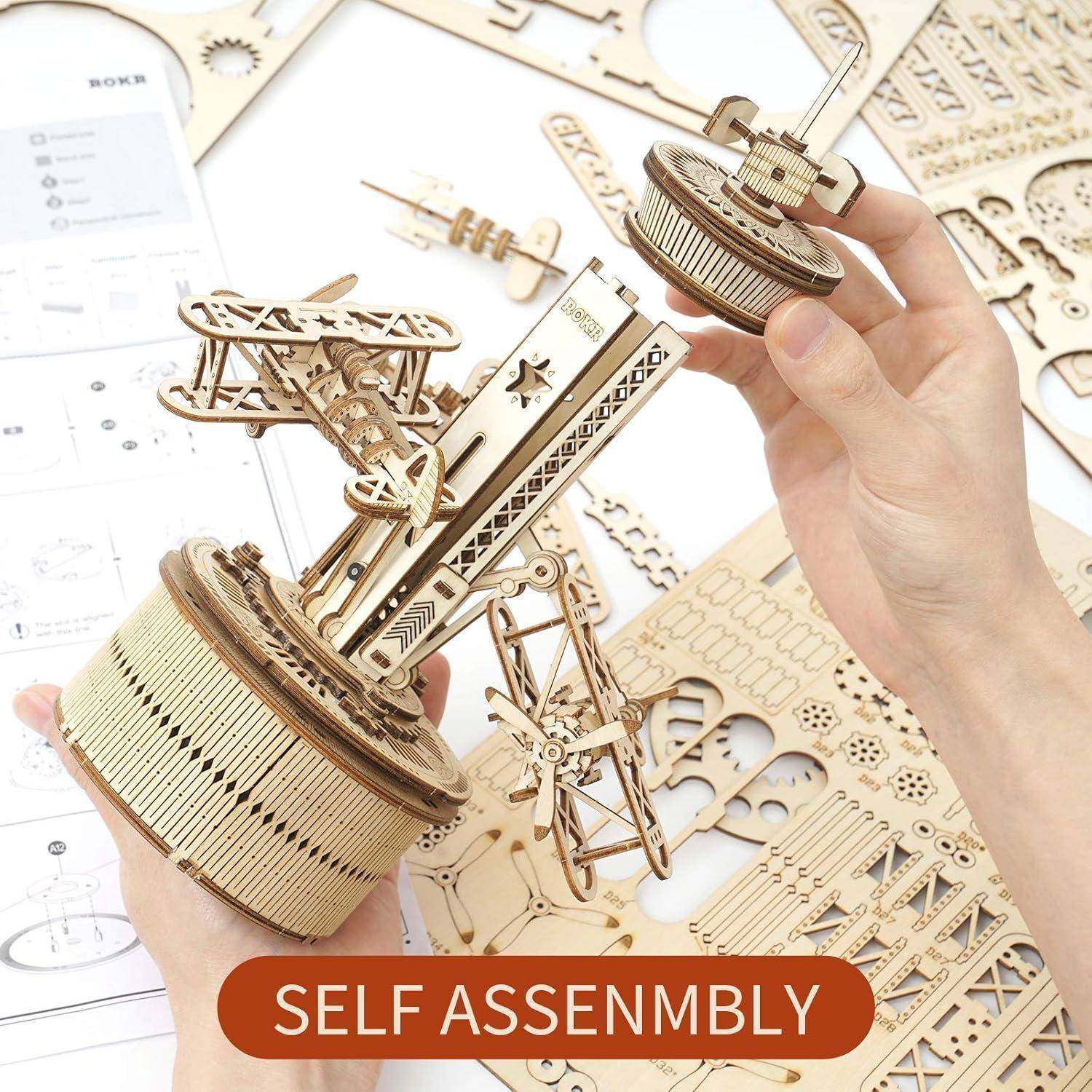 Zwei Hände, die ein hölzernes mechanisches Modell aus einem Bausatz konstruieren, wobei filigrane Zahnräder und Teile sichtbar sind. Eine Beschriftung liest 'Selbstmontage'.