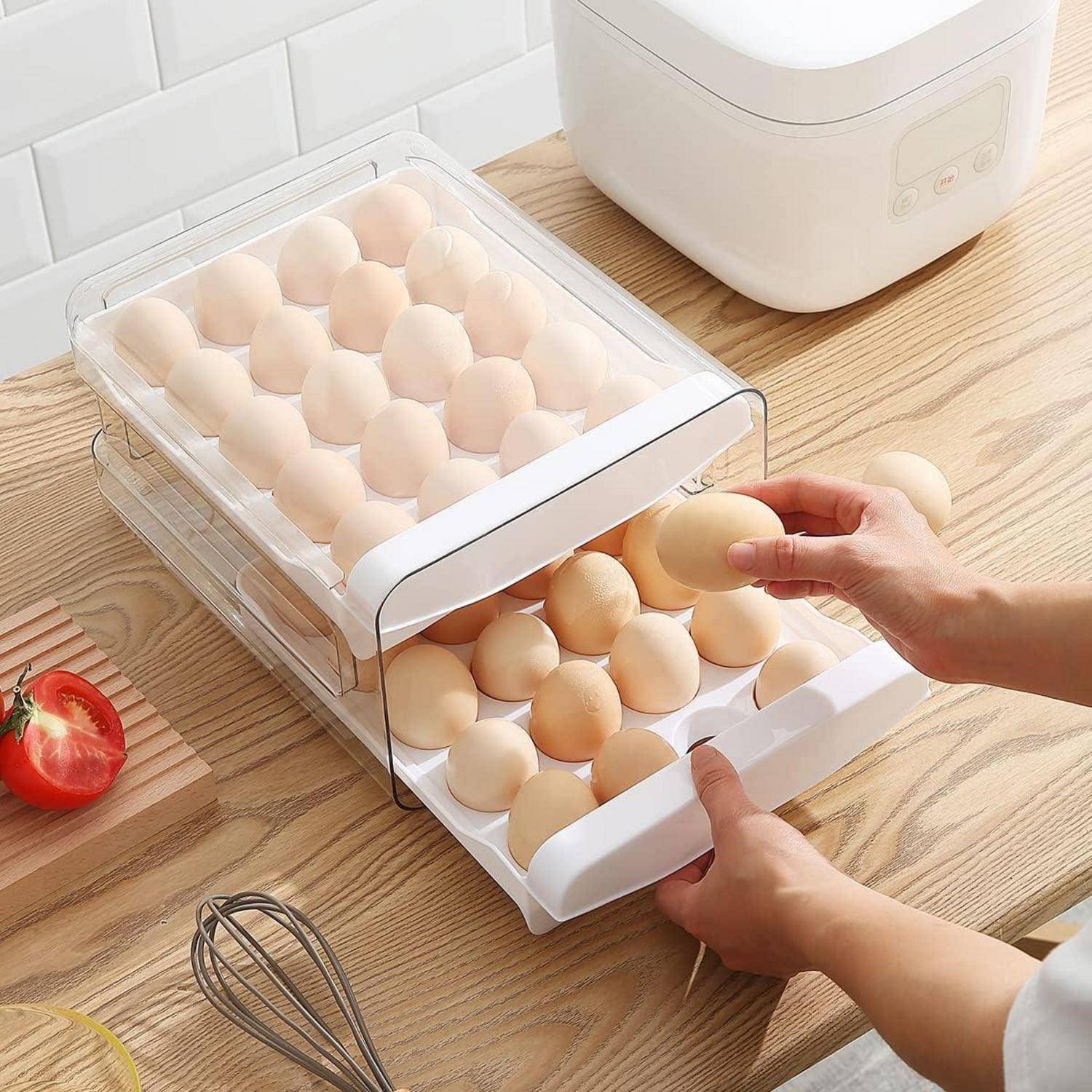 Hände sind zu sehen, die Eier in einem transparenten zweistöckigen Eierhalter auf einer Holzküchenzählplatte anordnen, neben einer Tomate, einem Schneebesen und einem Reiskocher.