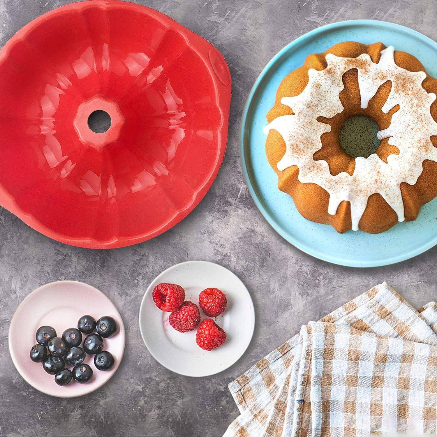 Ein Bundt-Kuchen mit weißer Glasur auf einem blauen Teller, daneben Blaubeeren, Erdbeeren und eine rote Form, auf einer grauen Oberfläche mit einem karierten Tuch.
