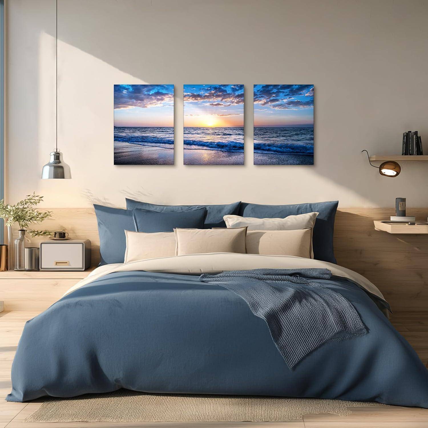 Ein gemütliches Schlafzimmer mit einem ordentlich bezogenen Bett in Blautönen, flankiert von zwei Nachttischen. Darüber hängt eine Triptychon-Darstellung eines Sonnenuntergangs über dem Meer.