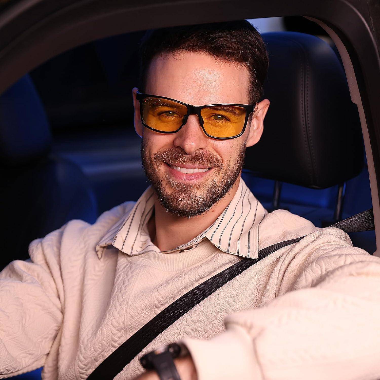 Ein Mann mit getönter Brille und einem cremefarbenen Pullover lächelt, während er in einem Auto sitzt.