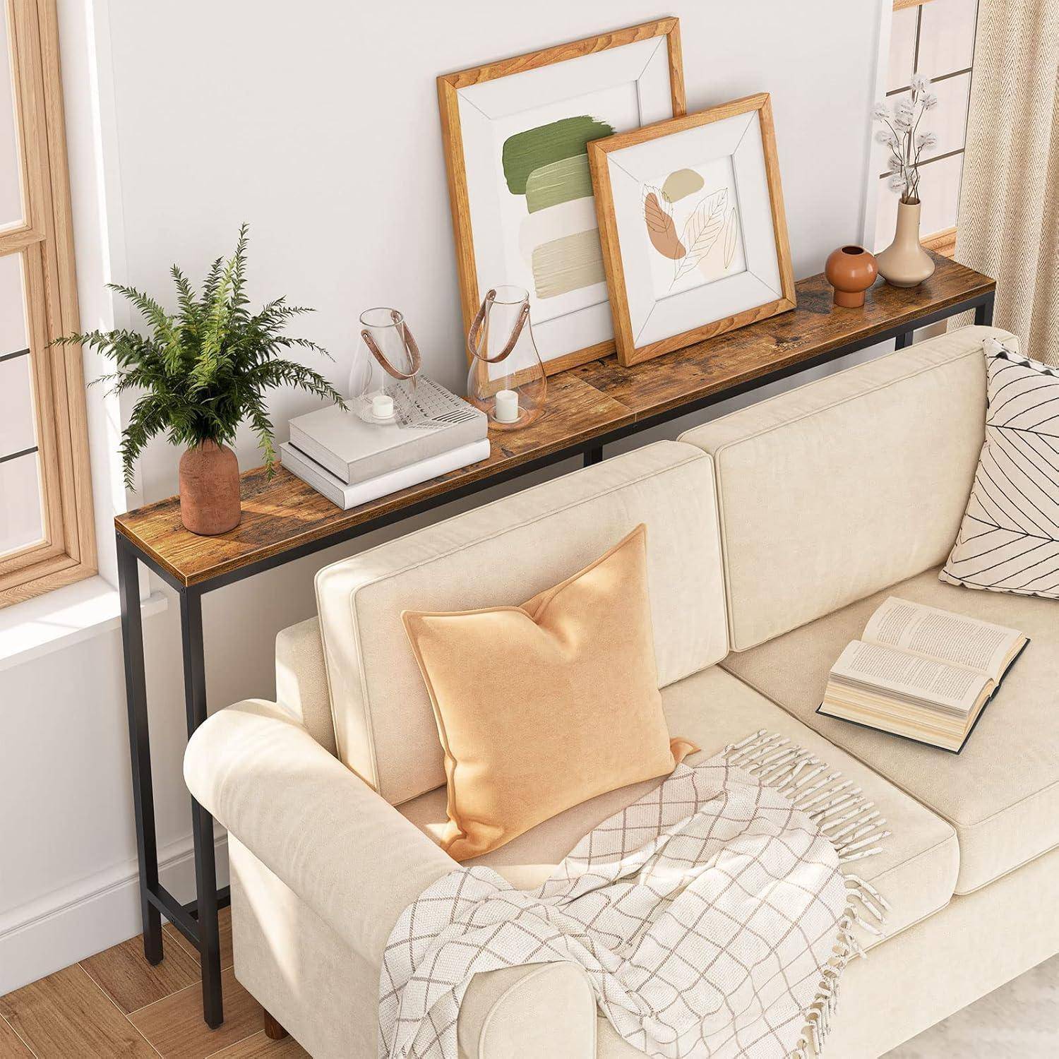 Eine gemütliche Ecke des Wohnzimmers mit einem beigen Sofa, gemusterter Decke und Kissen. Hinter dem Sofa steht ein Holzkonsoltisch, der Pflanzen, Bücher und gerahmte Kunstwerke trägt.