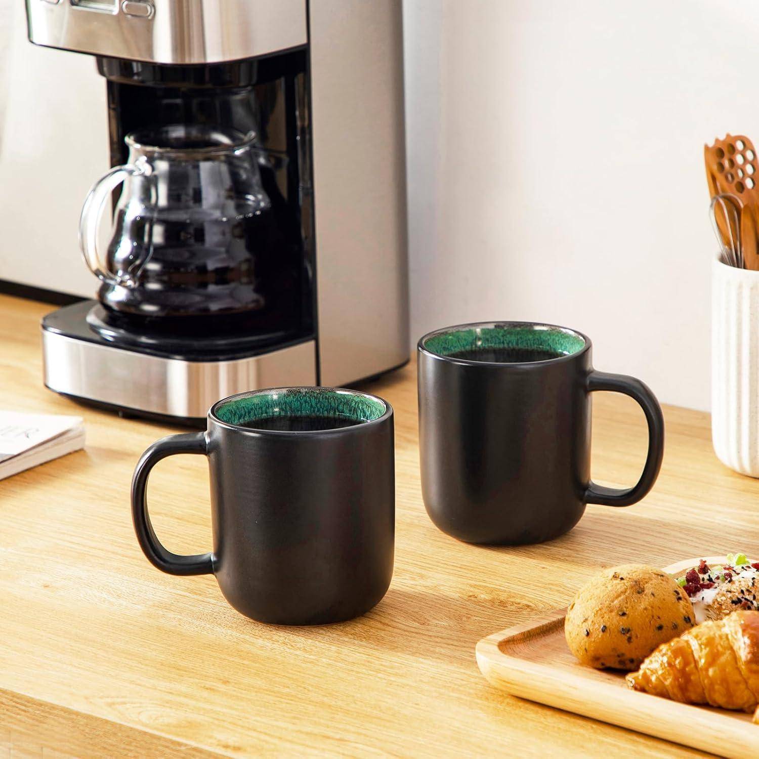 Eine Kaffeemaschine mit zwei schwarzen Tassen gefüllt mit Kaffee neben einem Holztablett mit Gebäckstücken auf einer Küchenarbeitsplatte.