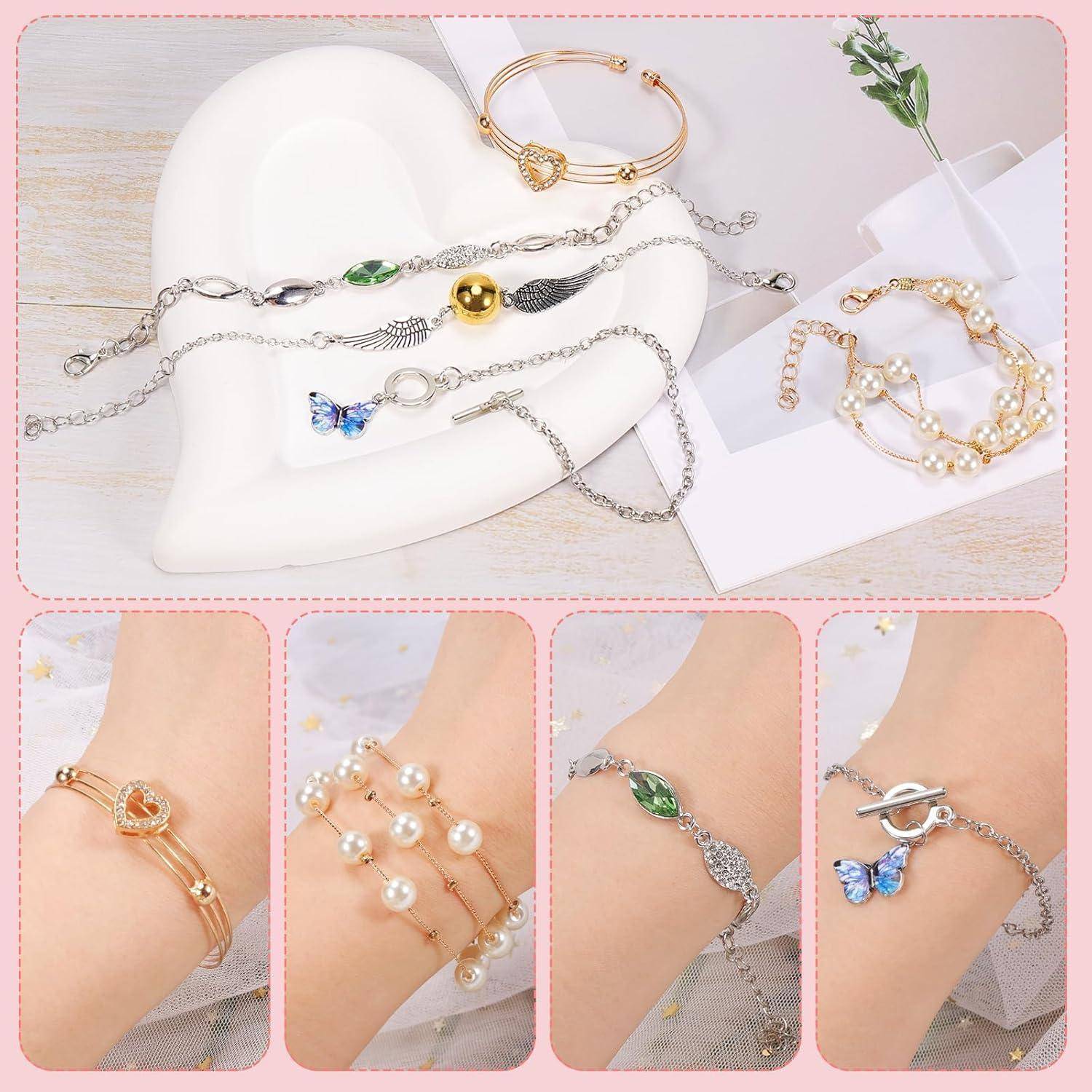 Eine Sammlung von Armbändern, die auf einer herzförmigen Schale ausgestellt und an Handgelenken getragen werden. Umfasst Gold-, Perlen- und Silberdesigns mit Anhängern.