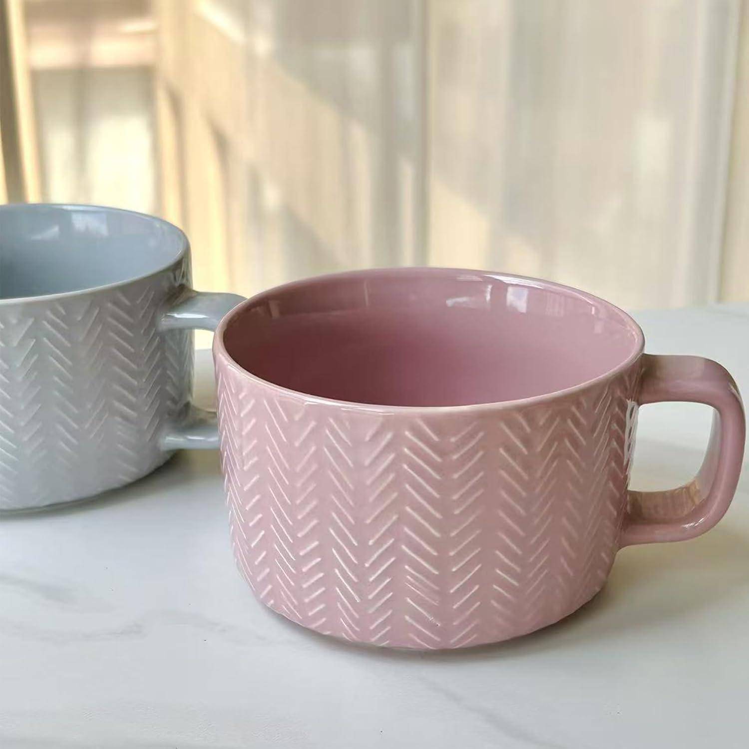 Zwei Keramikbecher mit Fischgrätenmuster, einer in Rosa und einer in Hellgrau, auf einer weißen Oberfläche in der Nähe von transparenten Gardinen.