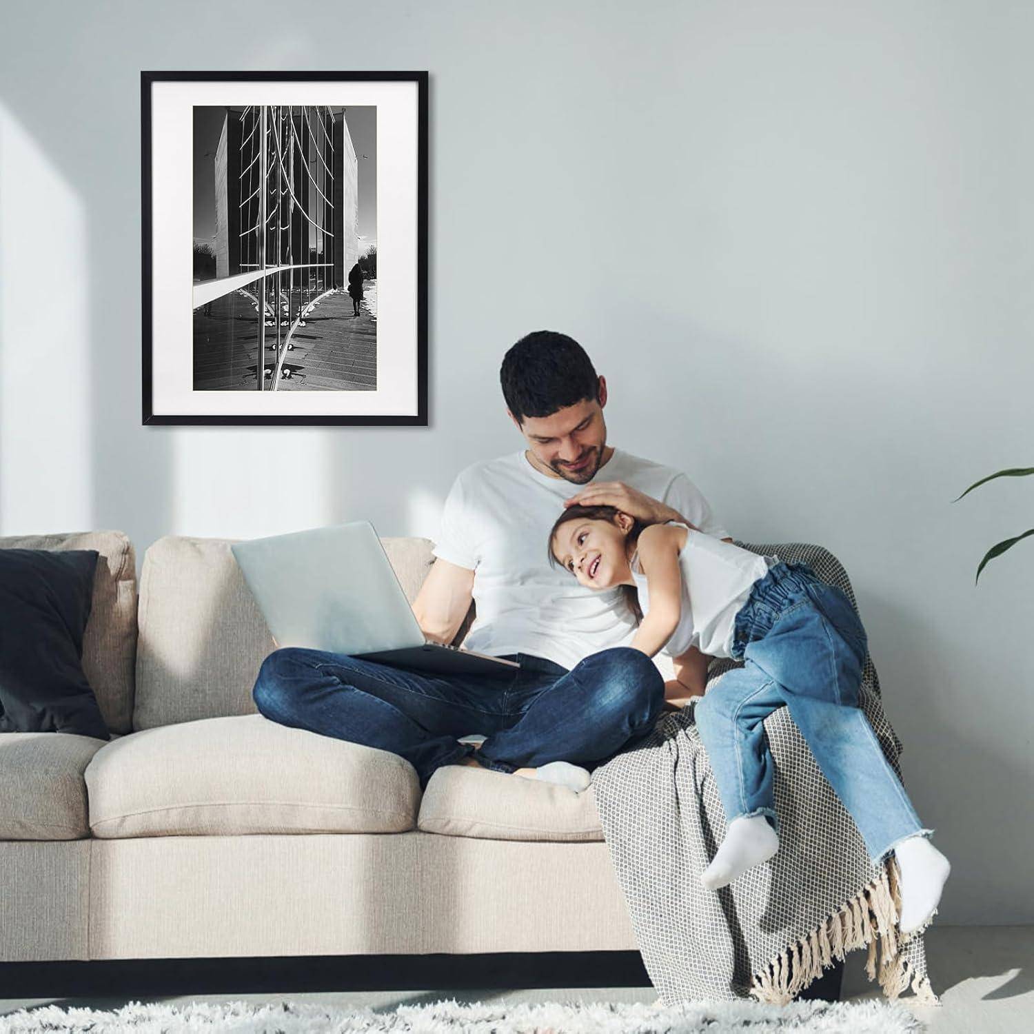 Ein Mann und ein junges Mädchen sitzen auf einem Sofa in einem hellen Wohnzimmer. Der Mann arbeitet an einem Laptop, während das Mädchen lächelt und sich an ihn lehnt. An der Wand hängt ein gerahmtes Foto.