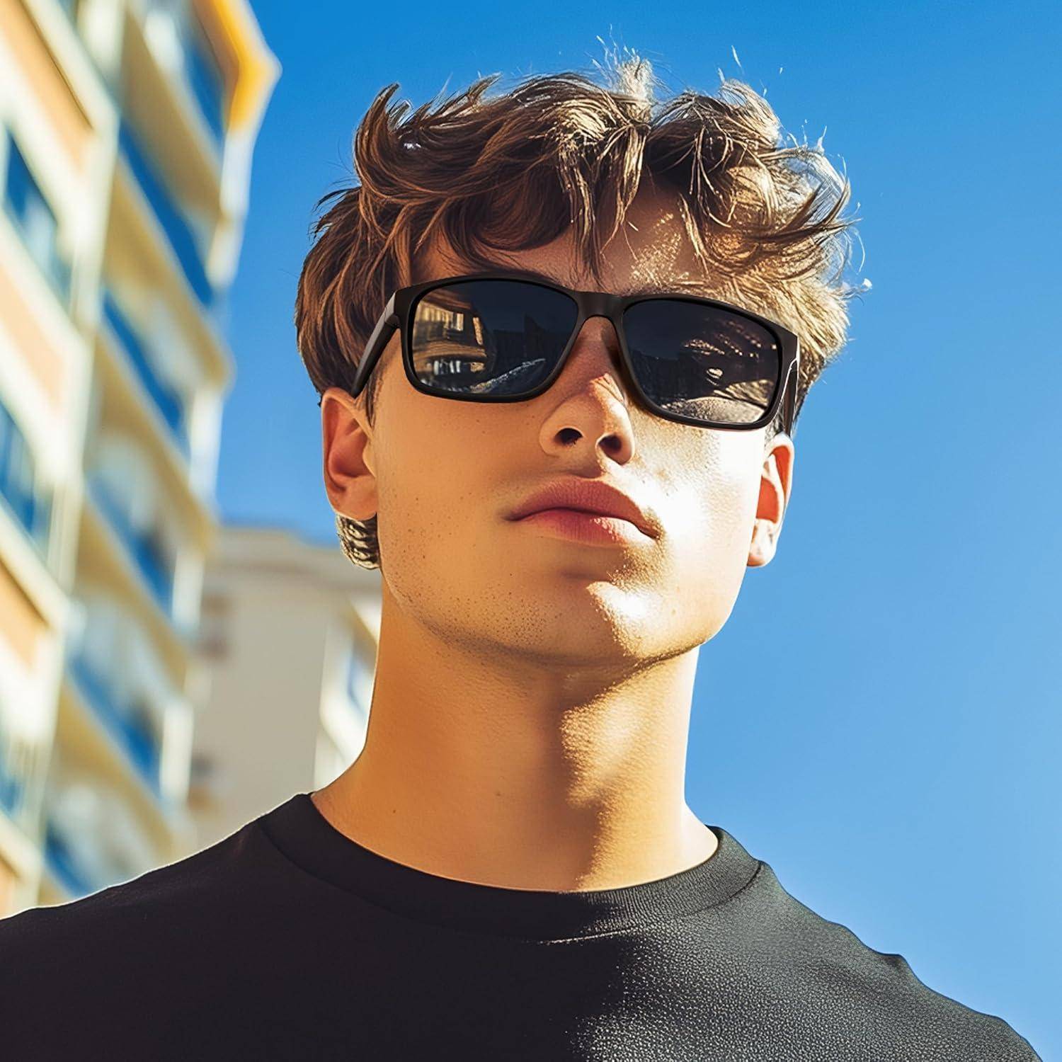 Eine junge Person mit Sonnenbrille und schwarzem Shirt steht draußen in der Nähe bunter Gebäude unter einem blauen Himmel und wirkt selbstbewusst.