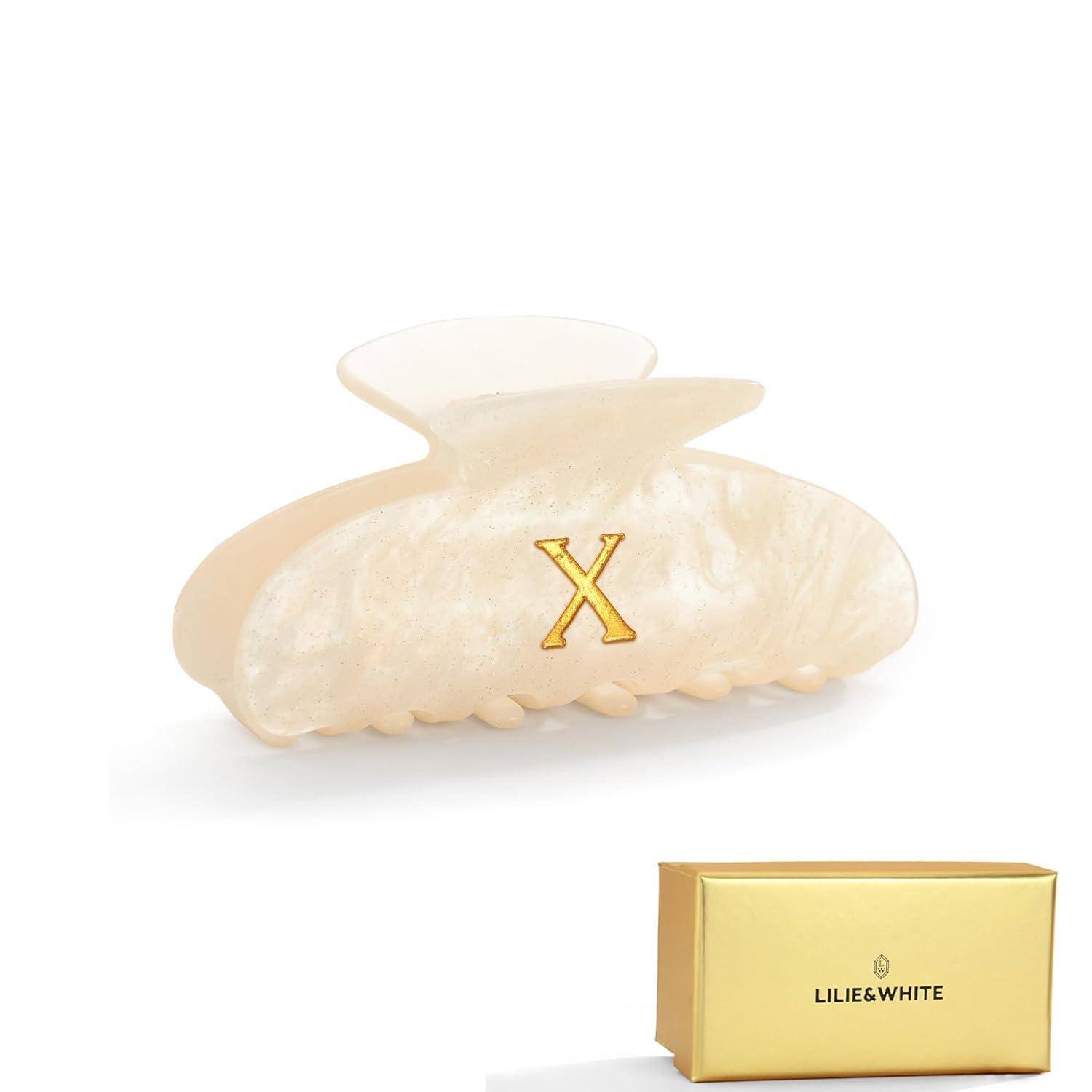 Eine perlweiße Haarspange mit einem goldenen 'X'-Logo, neben einer goldenen Schachtel mit der Aufschrift 'Lilie & White'.